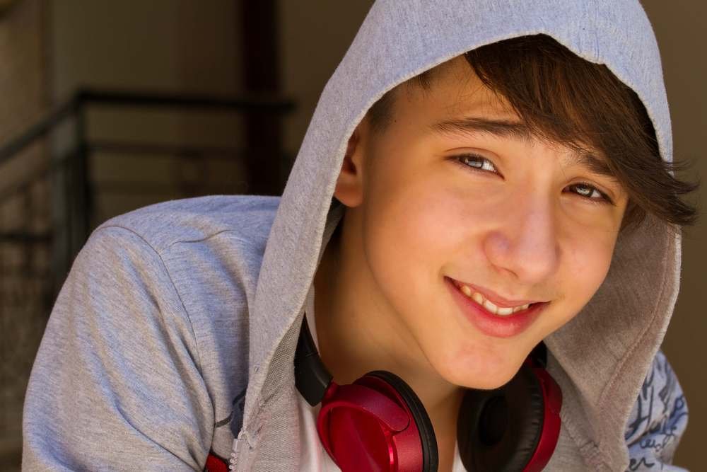 A teenage boy wearing a gray hoodie, with red headphones around his neck, smiling at the camera.