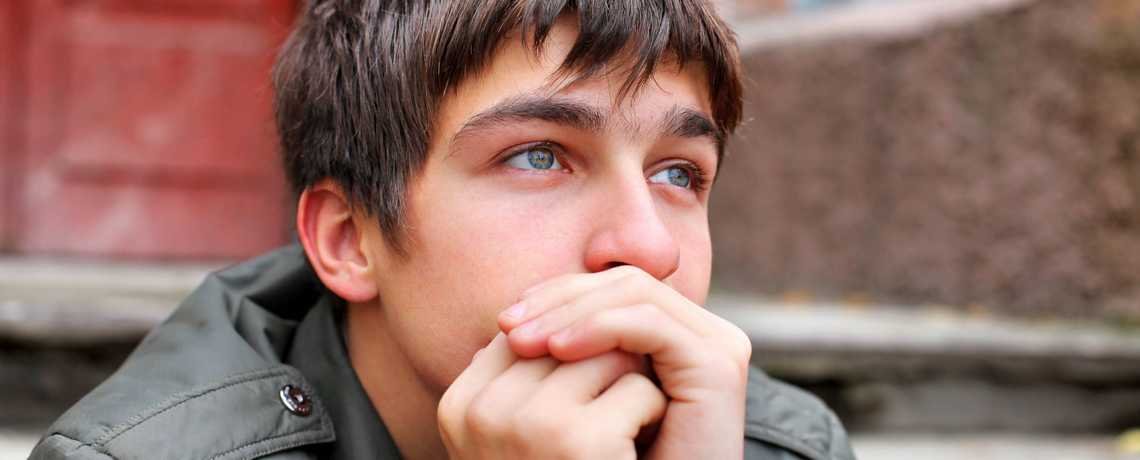 A young man with brown hair and blue eyes, wearing a gray jacket, sitting outdoors near a brick building, with his hand covering his mouth and looking thoughtfully into the distance.