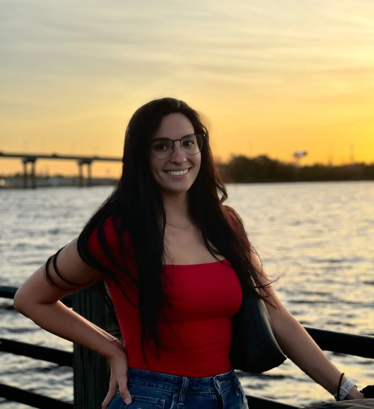 A young woman with dark hair and glasses smiling while standing near water at sunset, wearing a red top and blue jeans.