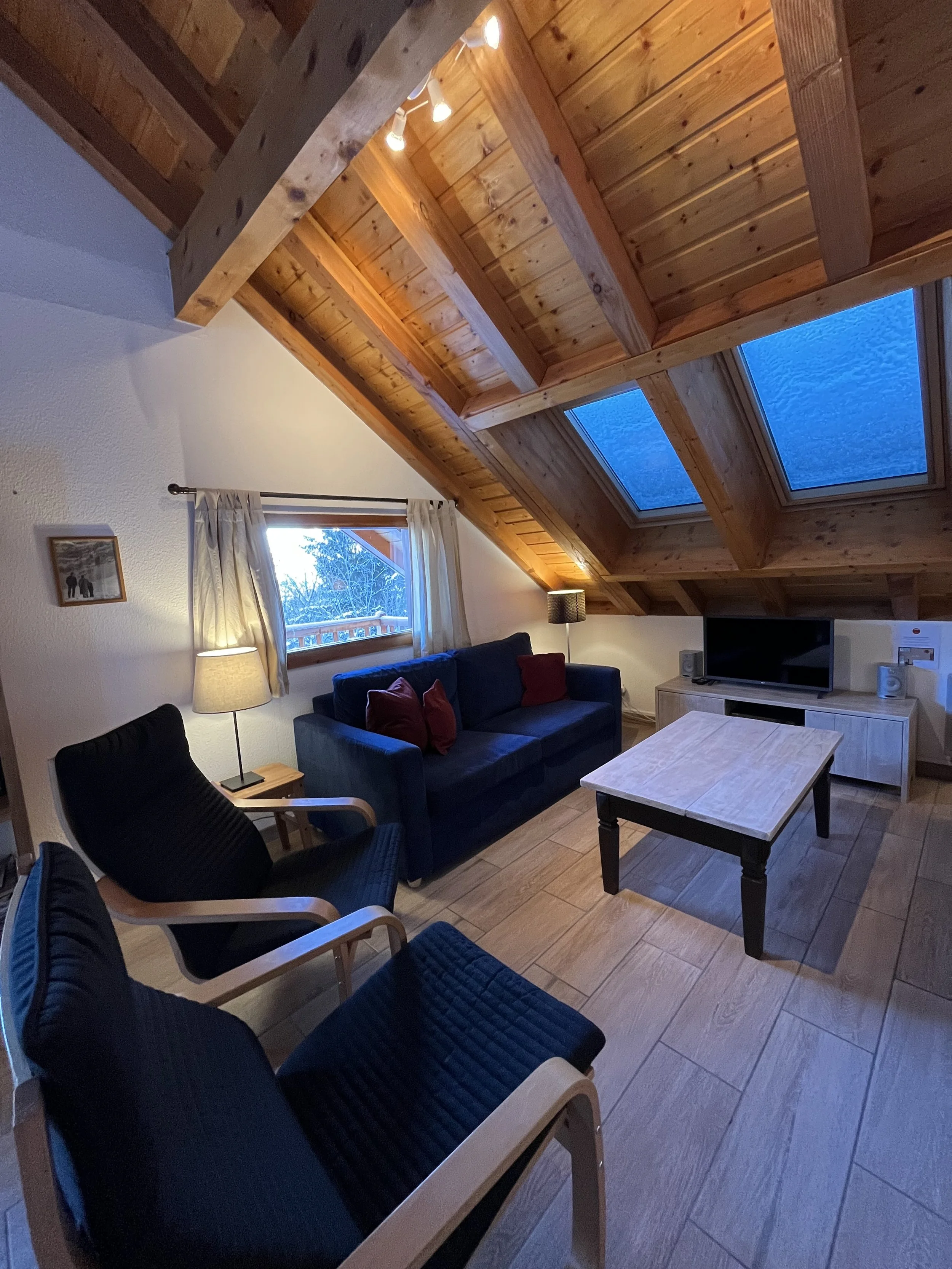 Cozy living room with a vaulted wooden ceiling, skylights, a window with curtains, a blue sofa with red pillows, two black armchairs with wooden frames, a wooden coffee table, a TV on a wooden stand, and warm lighting.