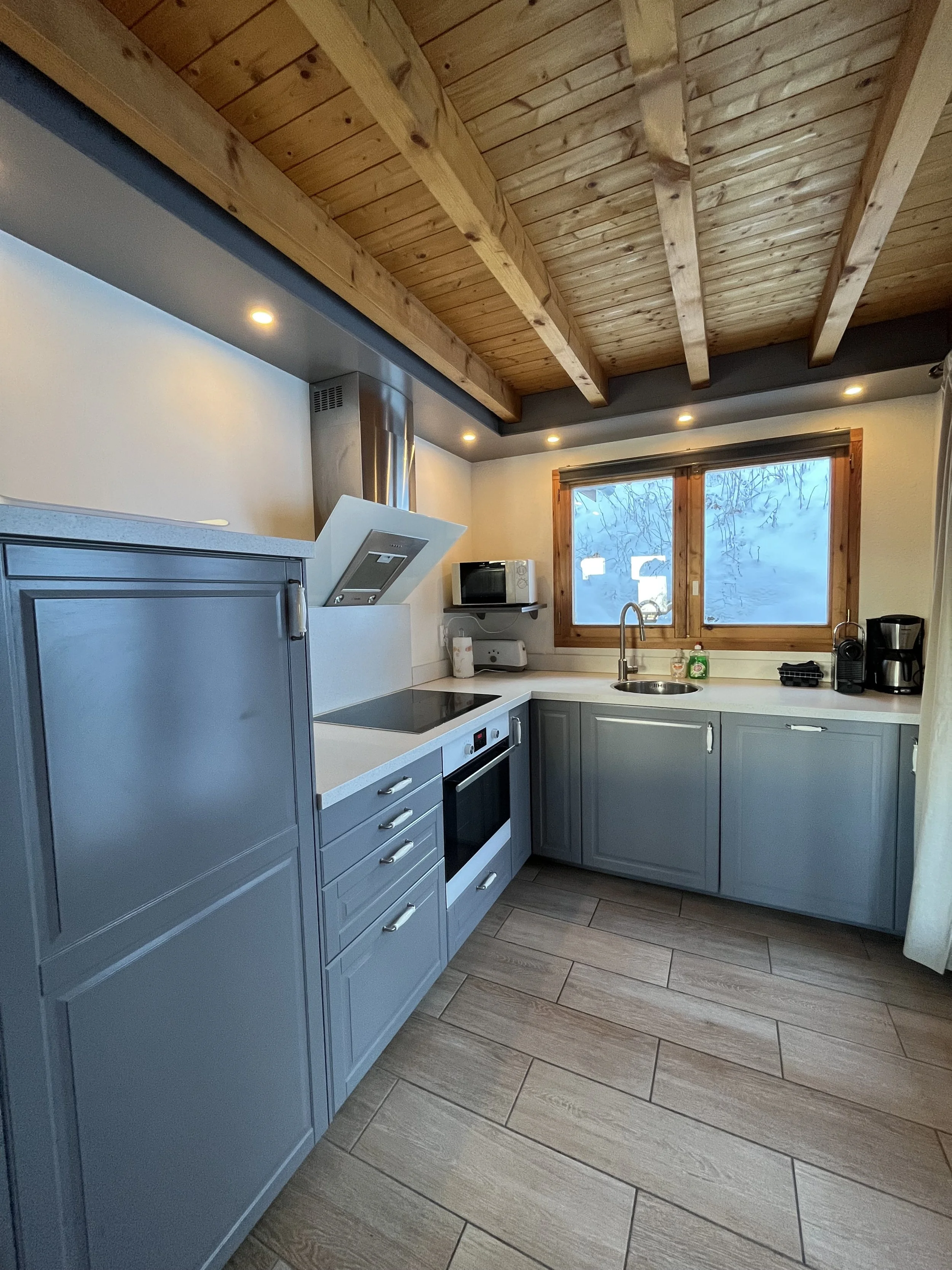 A compact kitchen with blue cabinets and white countertops, a stovetop, microwave, and coffee maker, under a wooden ceiling and next to a window showing a snowy landscape outside.