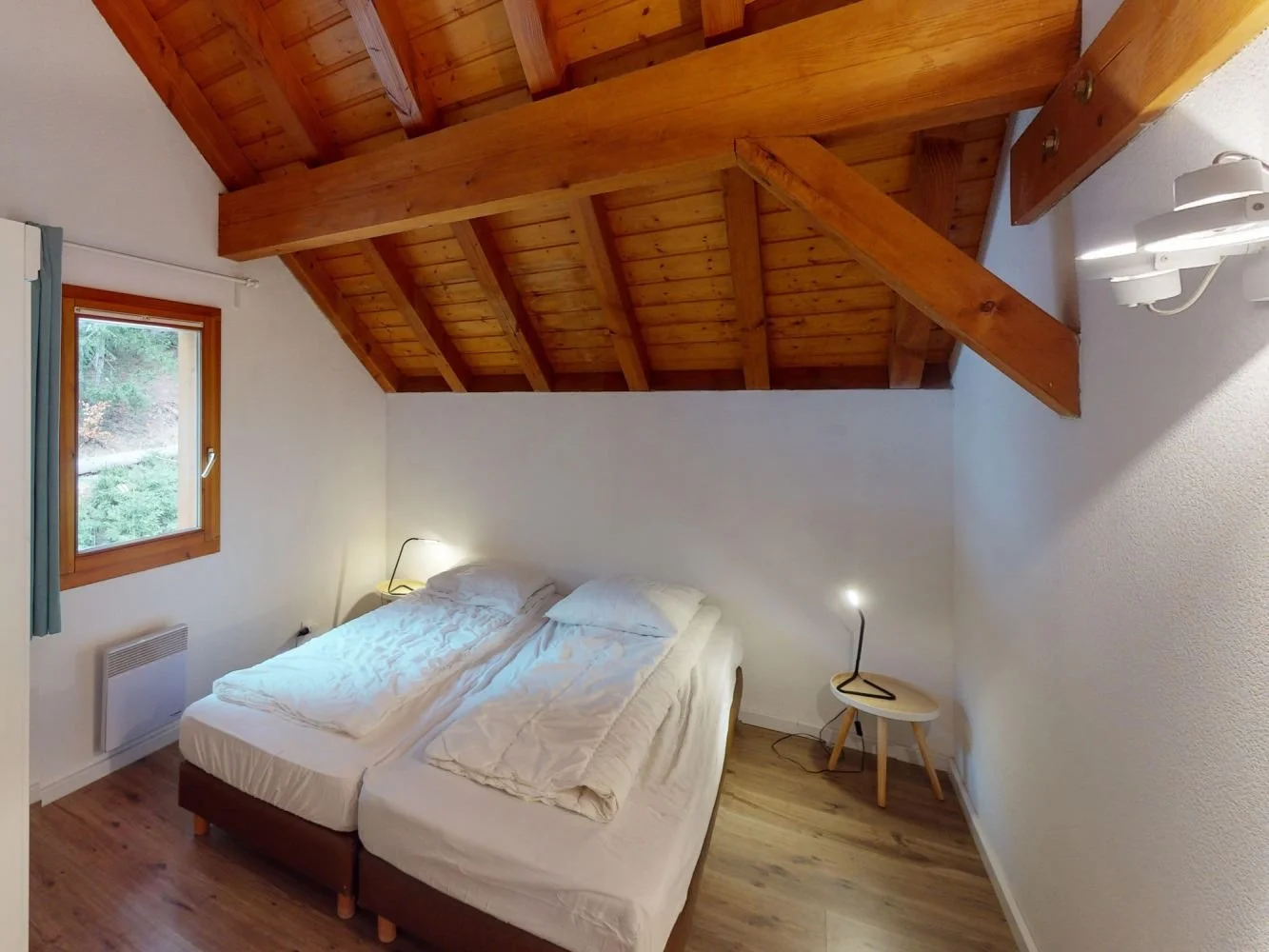 A bedroom with a wooden ceiling, a window with curtains, a bed with white bedding, two small bedside lamps, and a small table.