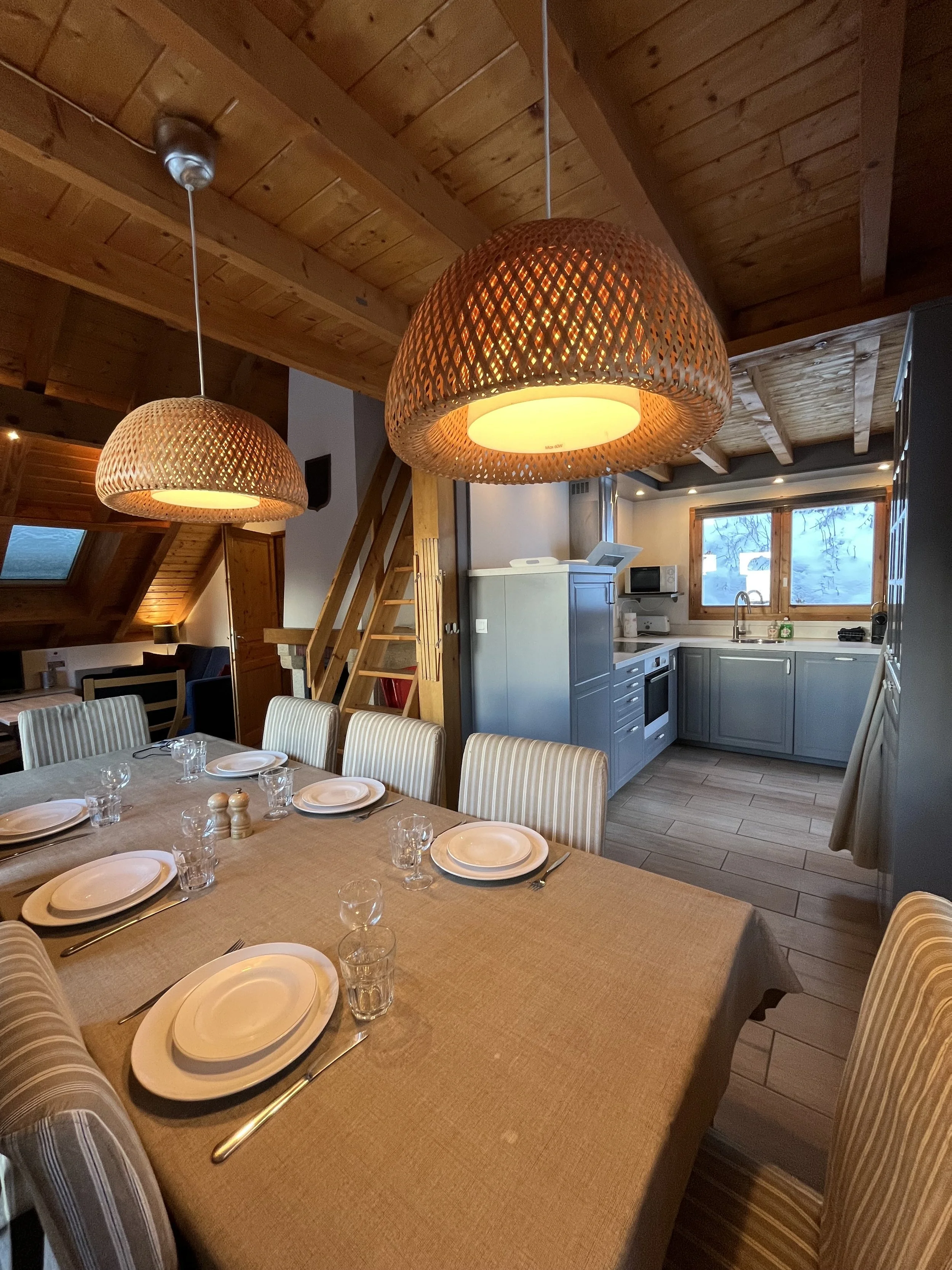 A cozy dining area with a large table set with plates, glasses, and cutlery, in front of a kitchen with blue cabinetry, a window, and wooden beams on the ceiling, illuminated by hanging woven pendant lights.