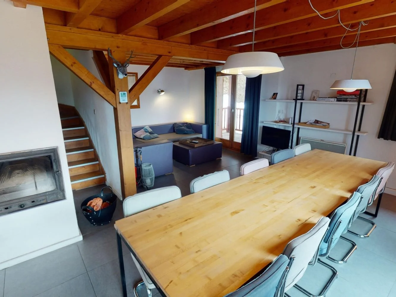 A spacious, open-concept living and dining area with a large wooden dining table surrounded by white chairs, a seating area with a gray sofa and pillows, a TV on a stand, a tall shelving unit with books and decor, and wooden ceiling beams with two ha