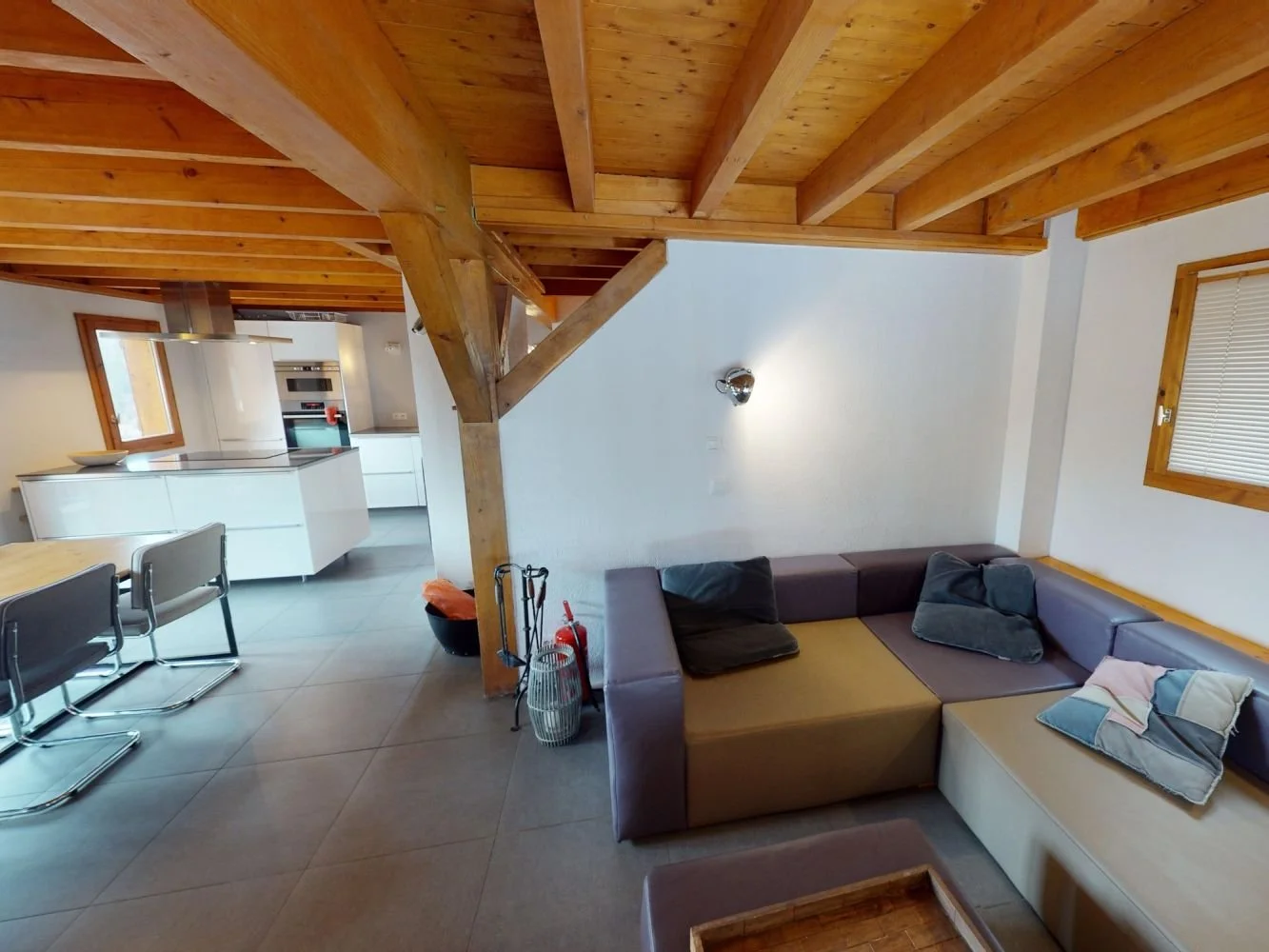 Living room with a sectional sofa with pillows, wood ceiling beams, and a next to a window with blinds. The open kitchen has white cabinetry, a stove, and a dining table with chairs.