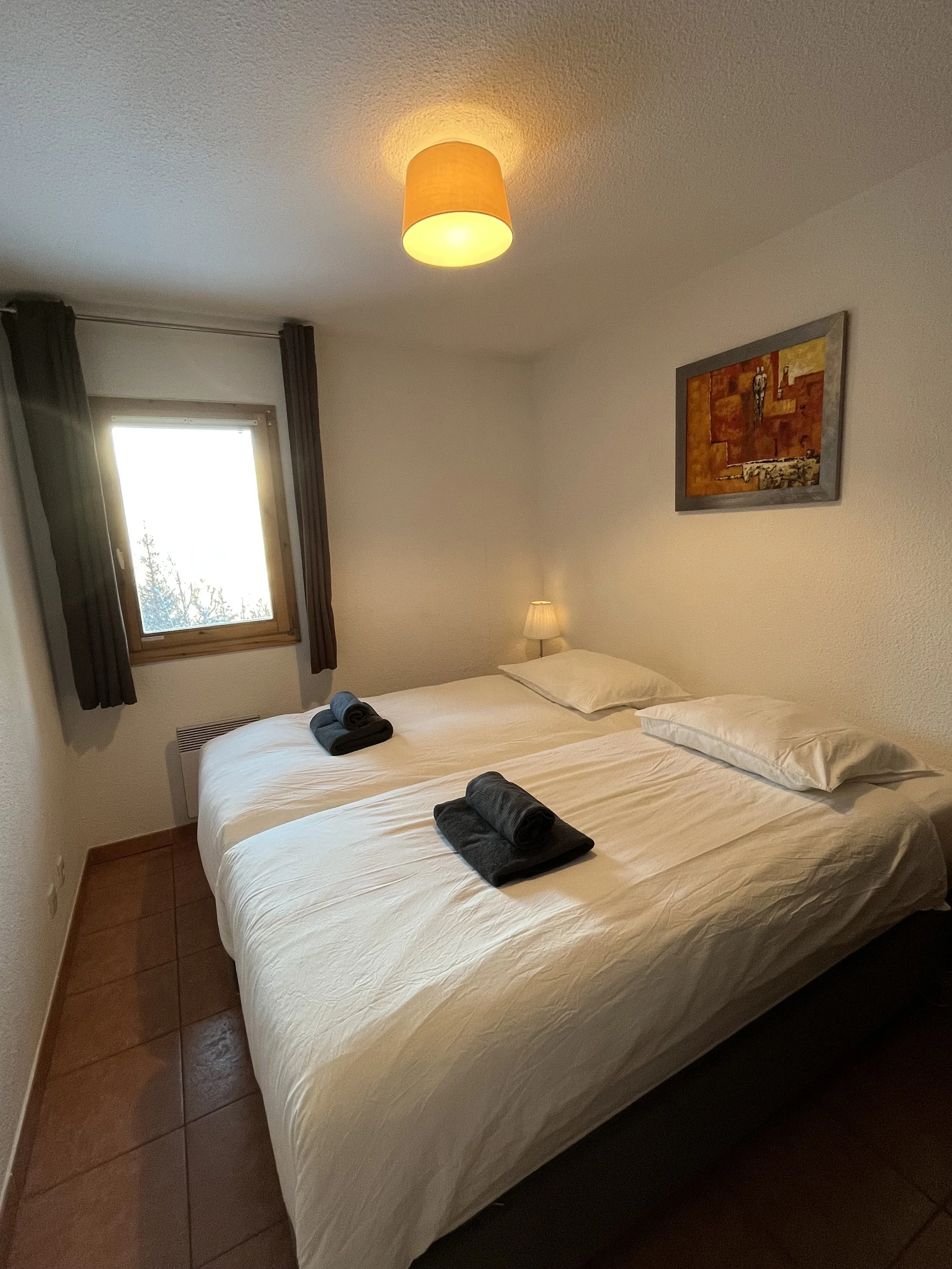 A cozy hotel bedroom featuring two twin beds with white linens, each topped with a rolled black towel. The room has a small window with dark curtains, a wall-mounted painting, a bedside lamp, and a ceiling light with an orange lampshade.