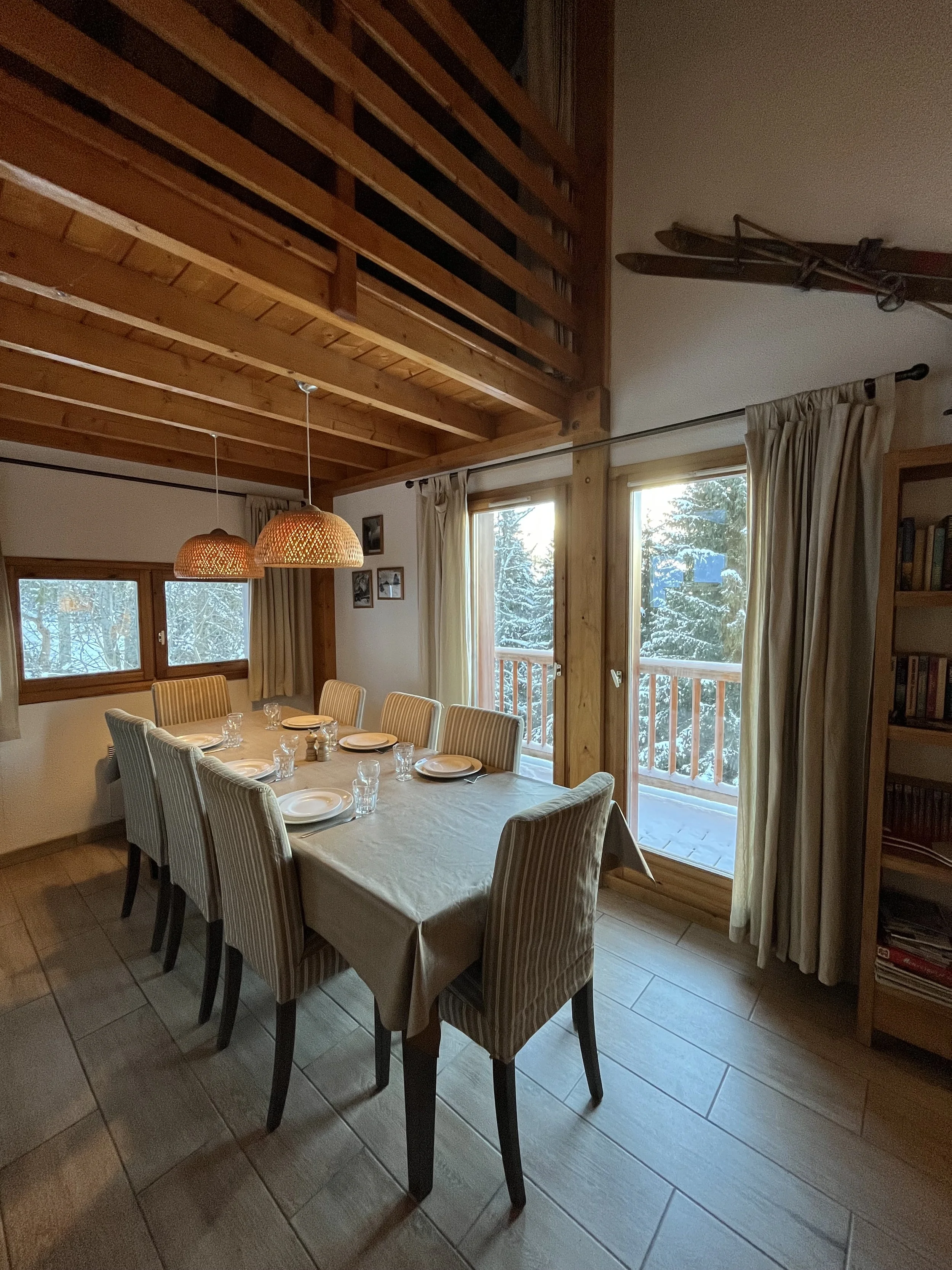 Dining room with a table set with plates and glasses, large windows with curtains, wooden ceiling and railing, and a snow-covered outdoor view.
