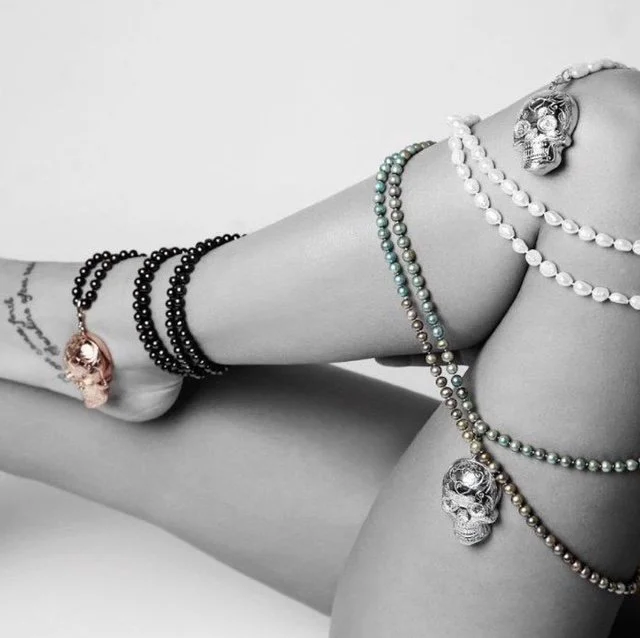 NYGP woman's leg with multiple pearl necklaces, bracelets, and skull-shaped jewellery in black, white, and metallic colours, against a plain background.