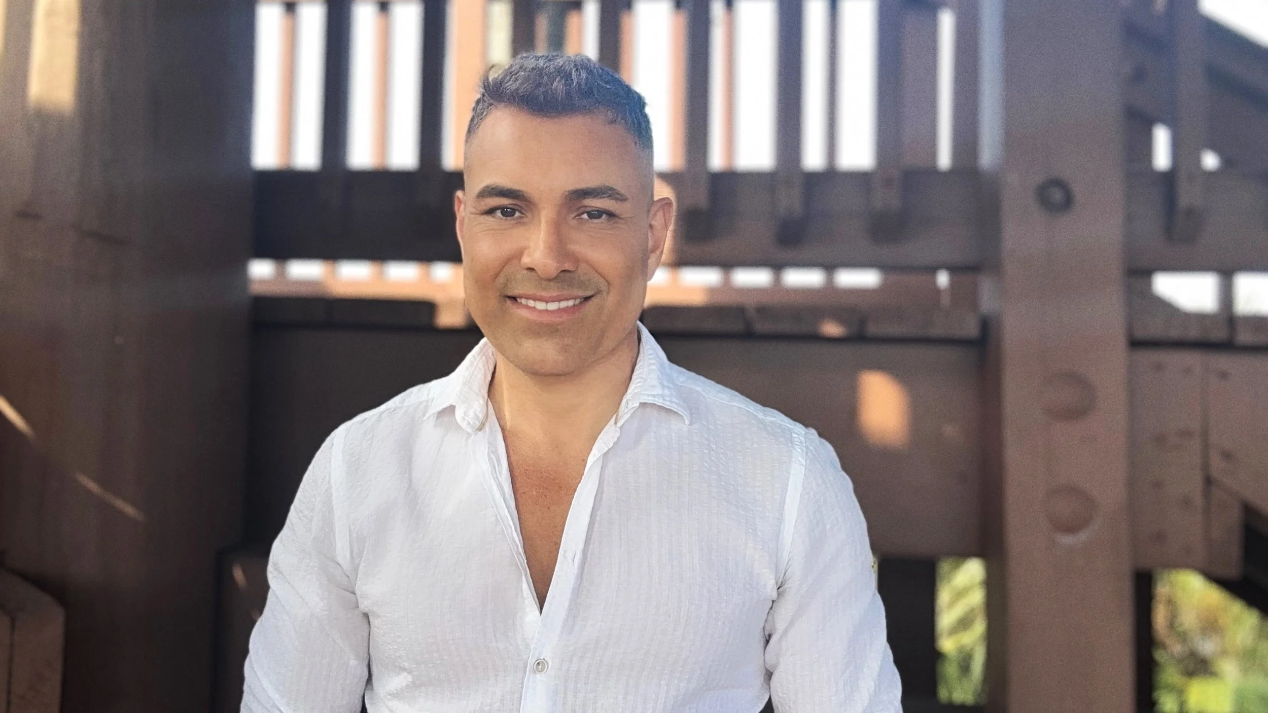 A smiling man with short dark hair wearing a white button-up shirt standing outdoors in front of a wooden structure with railing.