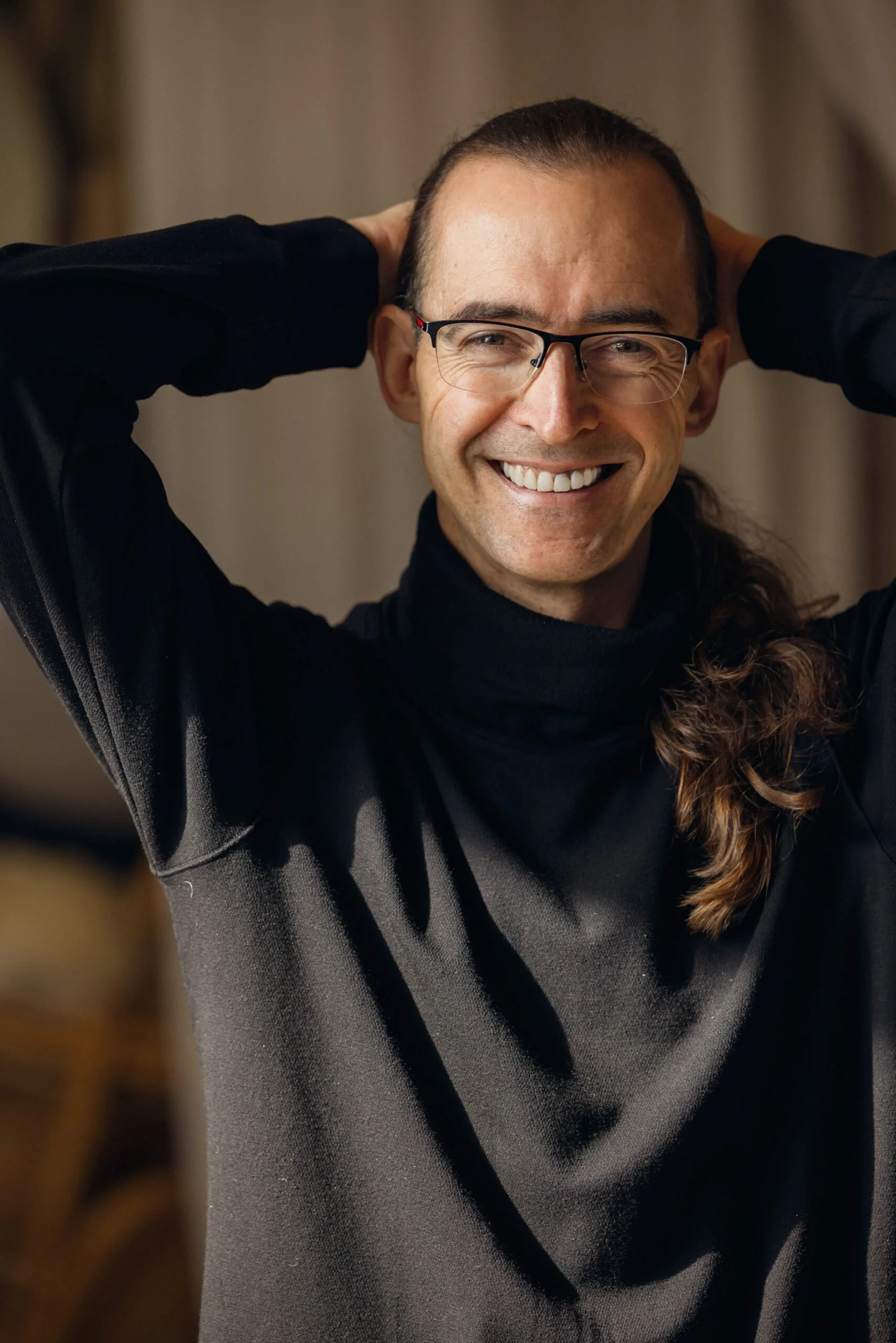 Ross Saunders smiling man with long hair and glasses, wearing a black long-sleeve shirt, with his hands behind his head in a warmly lit room.