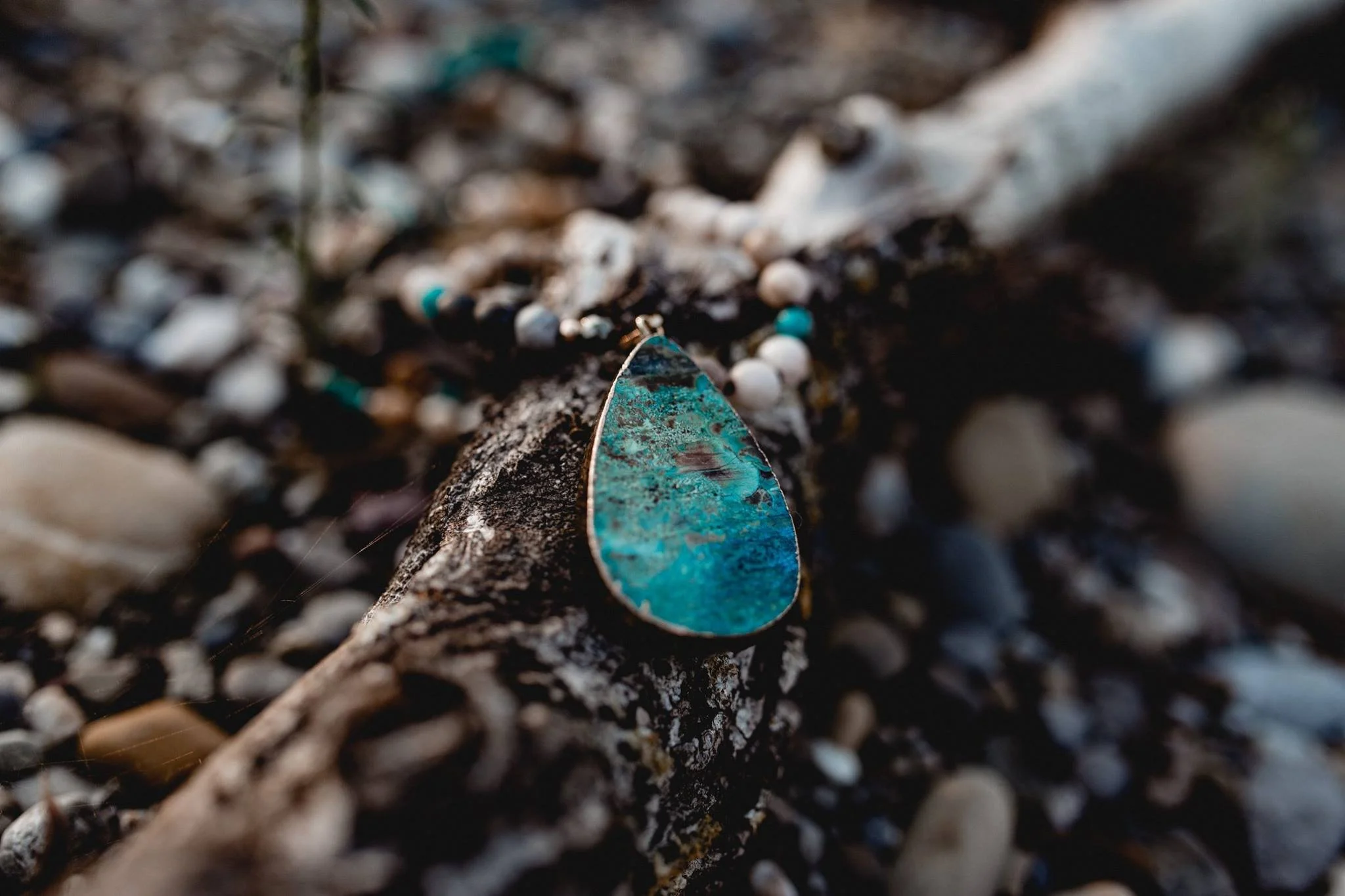 Ein türkisfarbener, tropfenförmiger Anhänger liegt auf einem Zweig mit kleinen Steinen und Sand. Die Umgebung ist natürlich und ruhig.