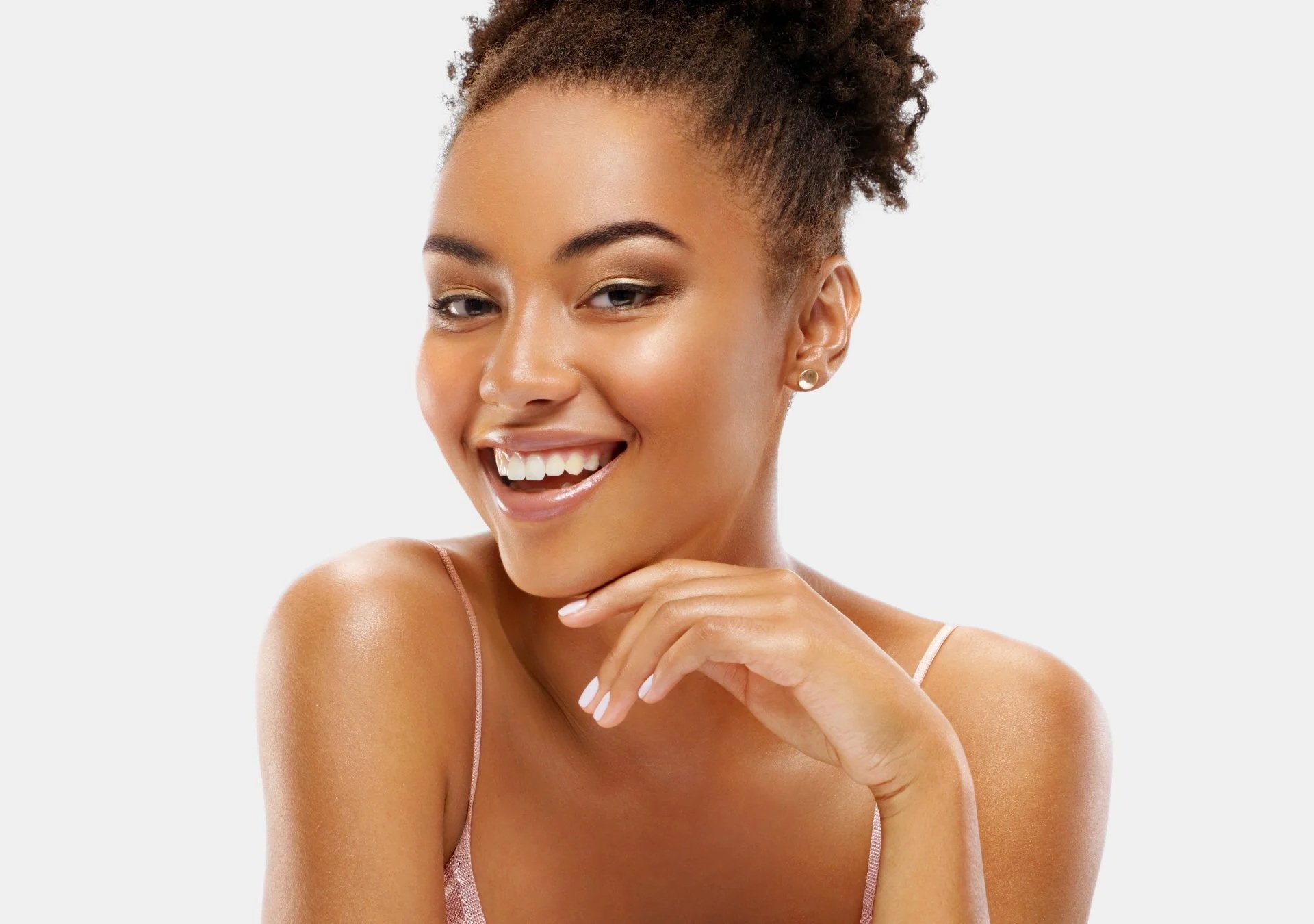 Jeune femme souriante avec peau éclatante, portant un haut à fines bretelles, posant avec la main sous le menton contre un fond blanc.