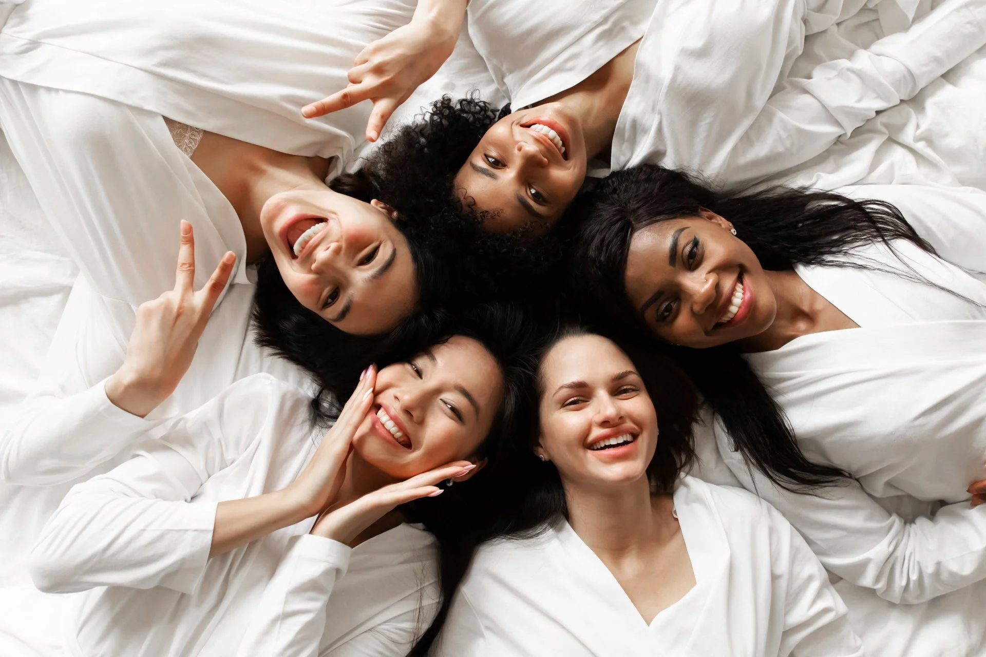 Cinq femmes de différents horizons ethniques allongées sur un lit blanc, souriantes et heureuses, se prenant en photo avec un téléphone pour un selfie.