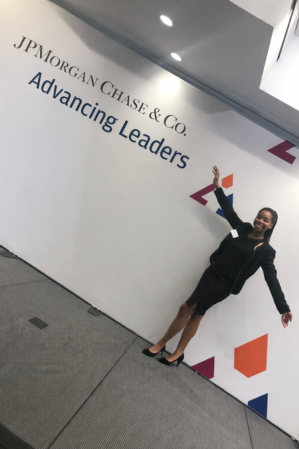 A woman in a black dress and blazer smiling and pointing at a sign that reads 'JPMorgan Chase & Co. Advancing Leaders'.