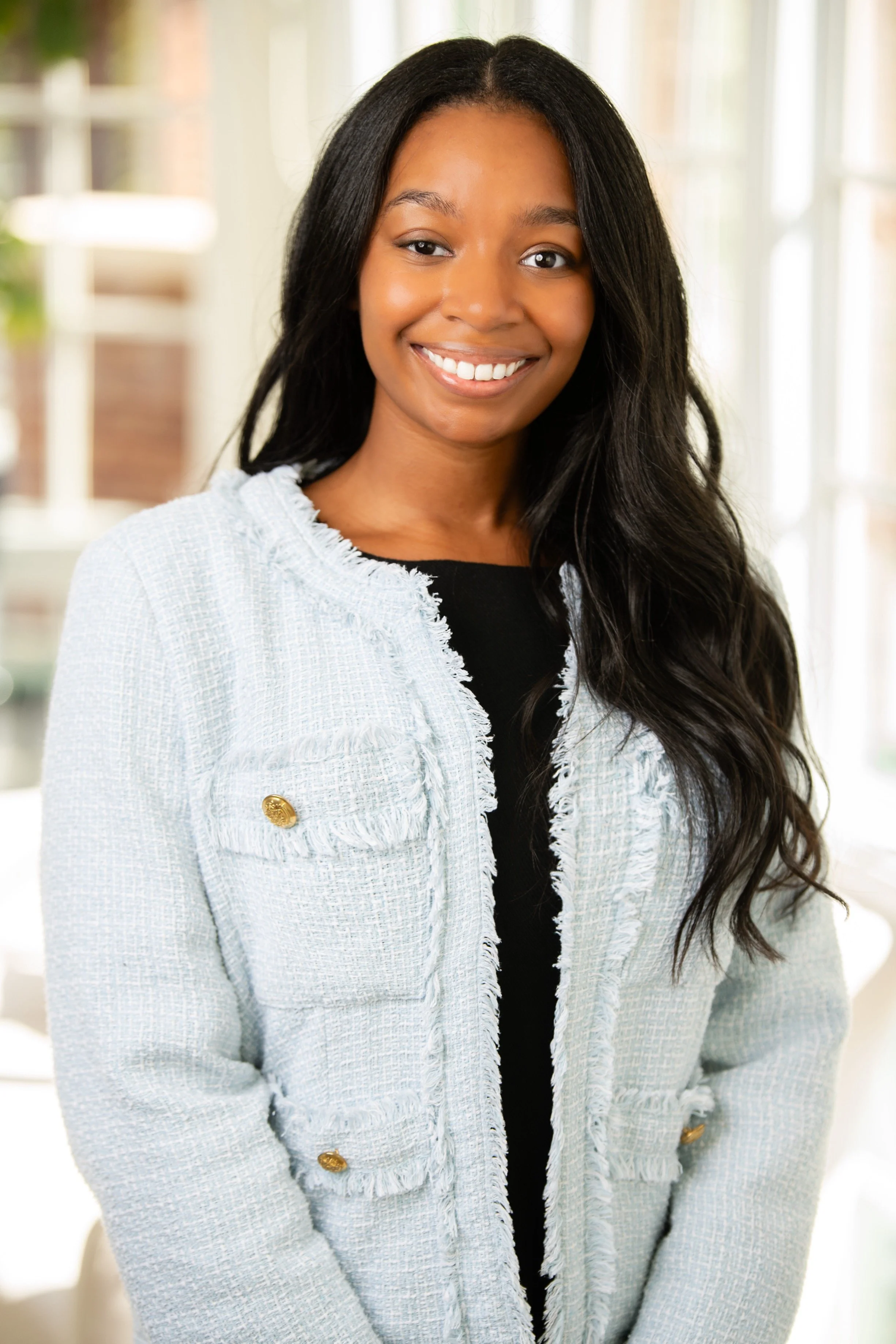 A woman with long black hair, smiling, wearing a light-colored textured blazer with gold buttons, standing indoors near windows.