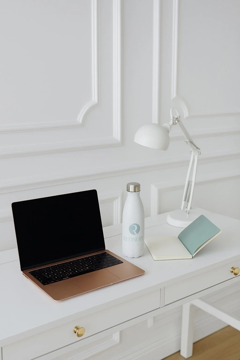A white desk with a rose gold laptop, a white water bottle labeled 'Refinery', a light blue open notebook, and a white adjustable desk lamp in a room with white walls and decorative molding.