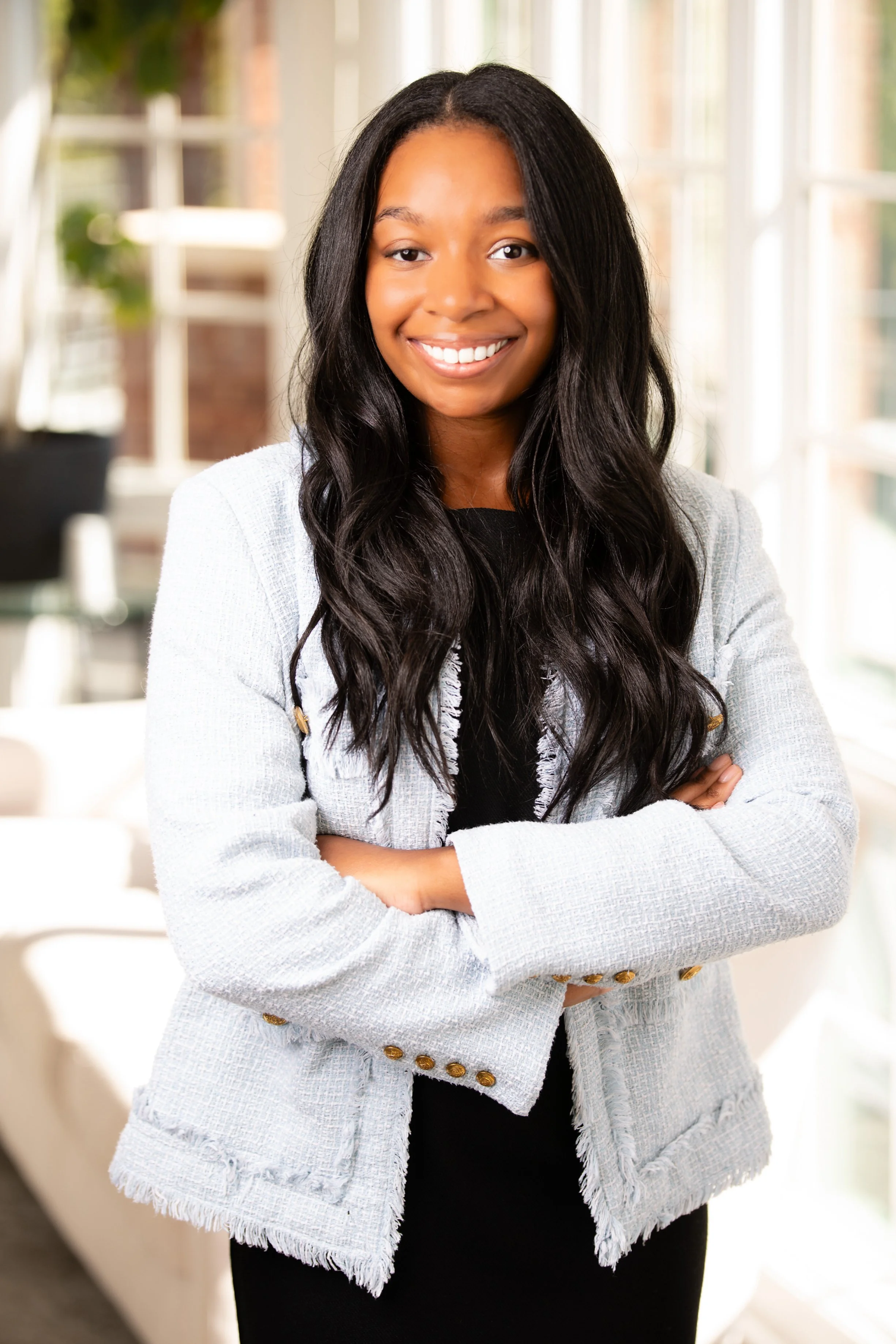 Portrait of a young woman with long black wavy hair, smiling, standing indoors with arms crossed, wearing a light-colored blazer over a black top.