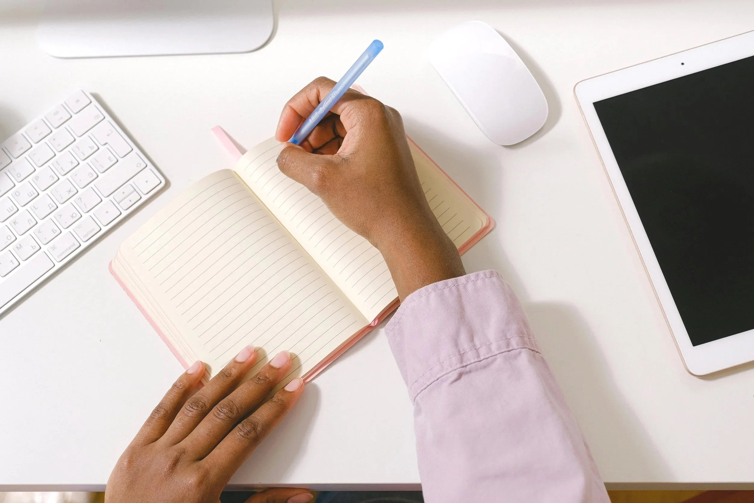 Person writing in a pink bordered notebook with a blue pen, surrounded by a white keyboard, a mouse, and a tablet on a white desk.