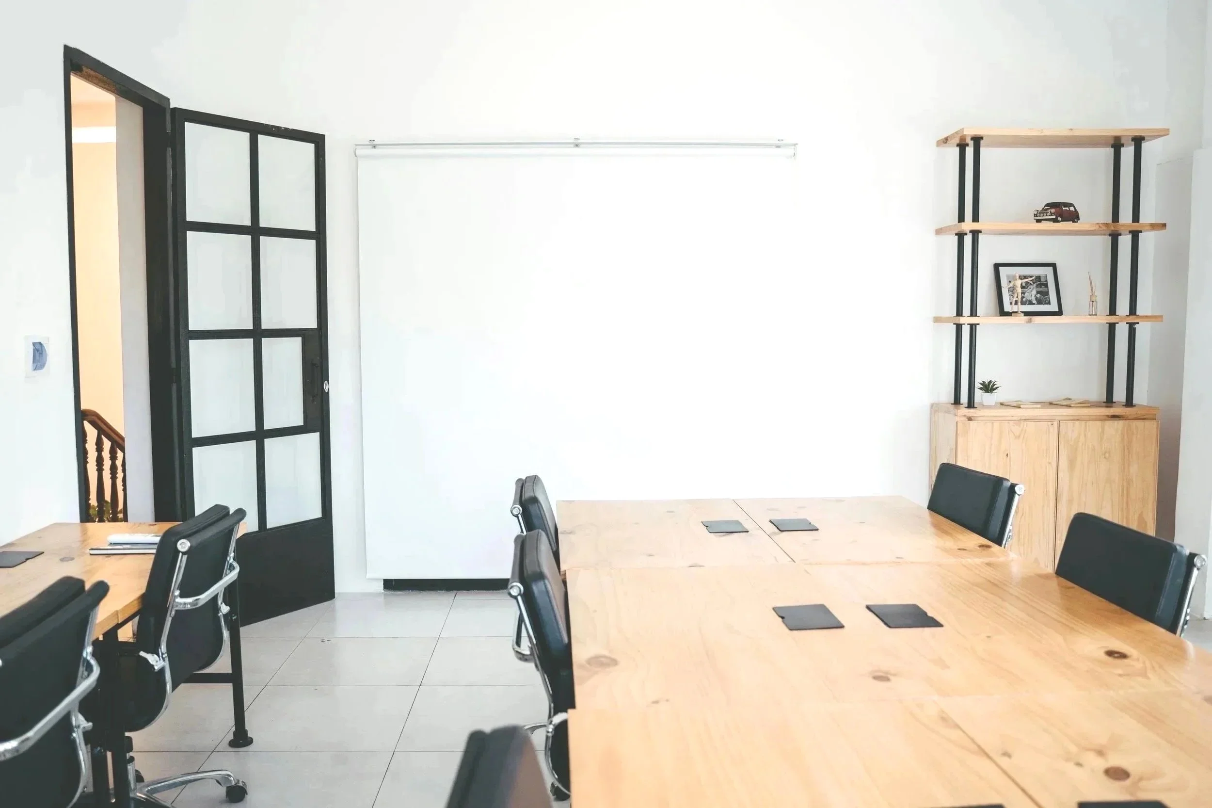 A modern conference room with a wooden table and black office chairs, a black-framed glass door, a white wall with a pull-down screen, and a wooden shelving unit with decorative items.