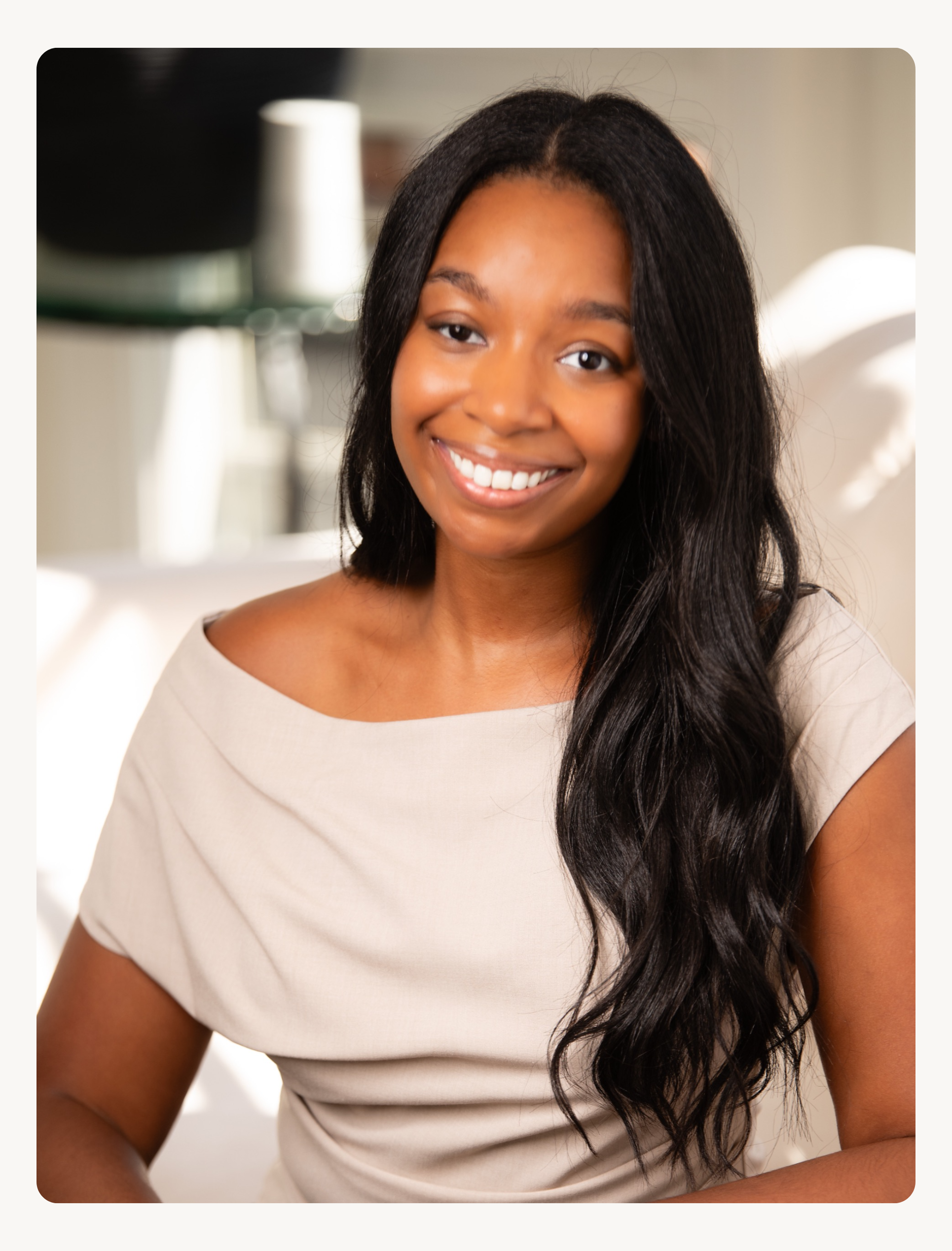 A woman with long black hair smiling while seated on a white couch in a well-lit room.