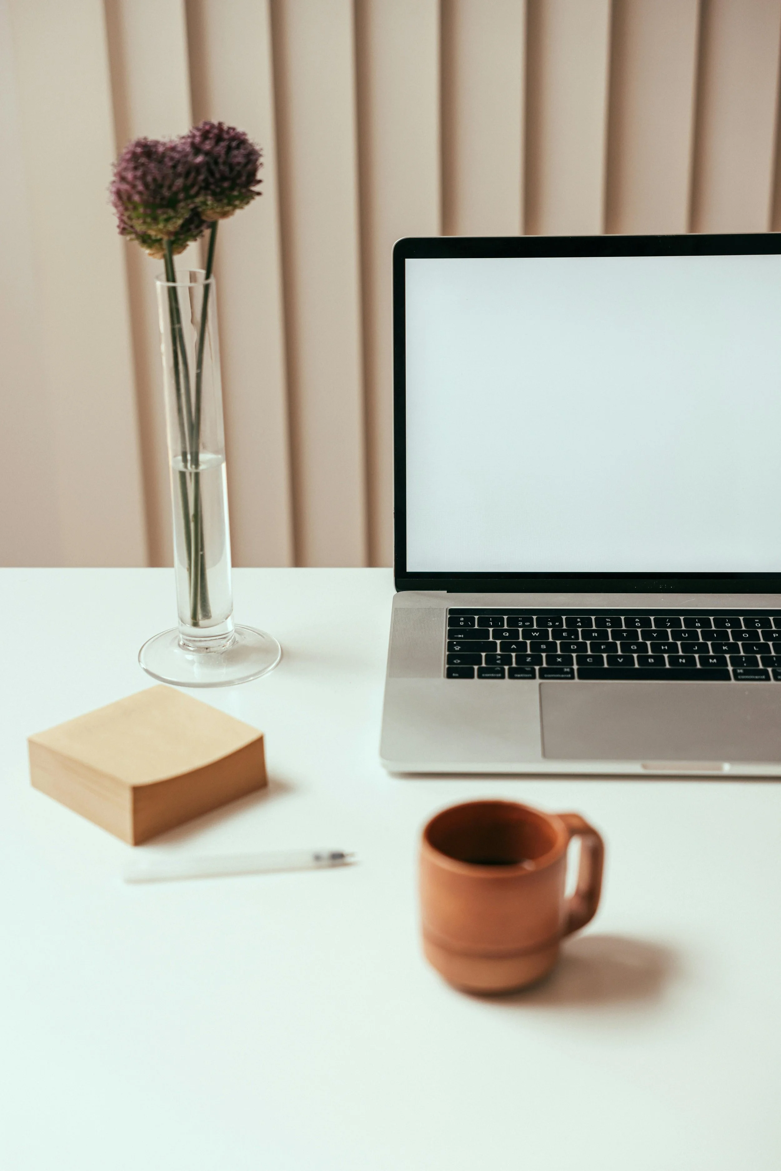 A minimalist workspace with a silver laptop, a brown mug, a glass vase with purple flowers, a wooden notepad, and a white pen on a white desk in front of a beige paneled wall.