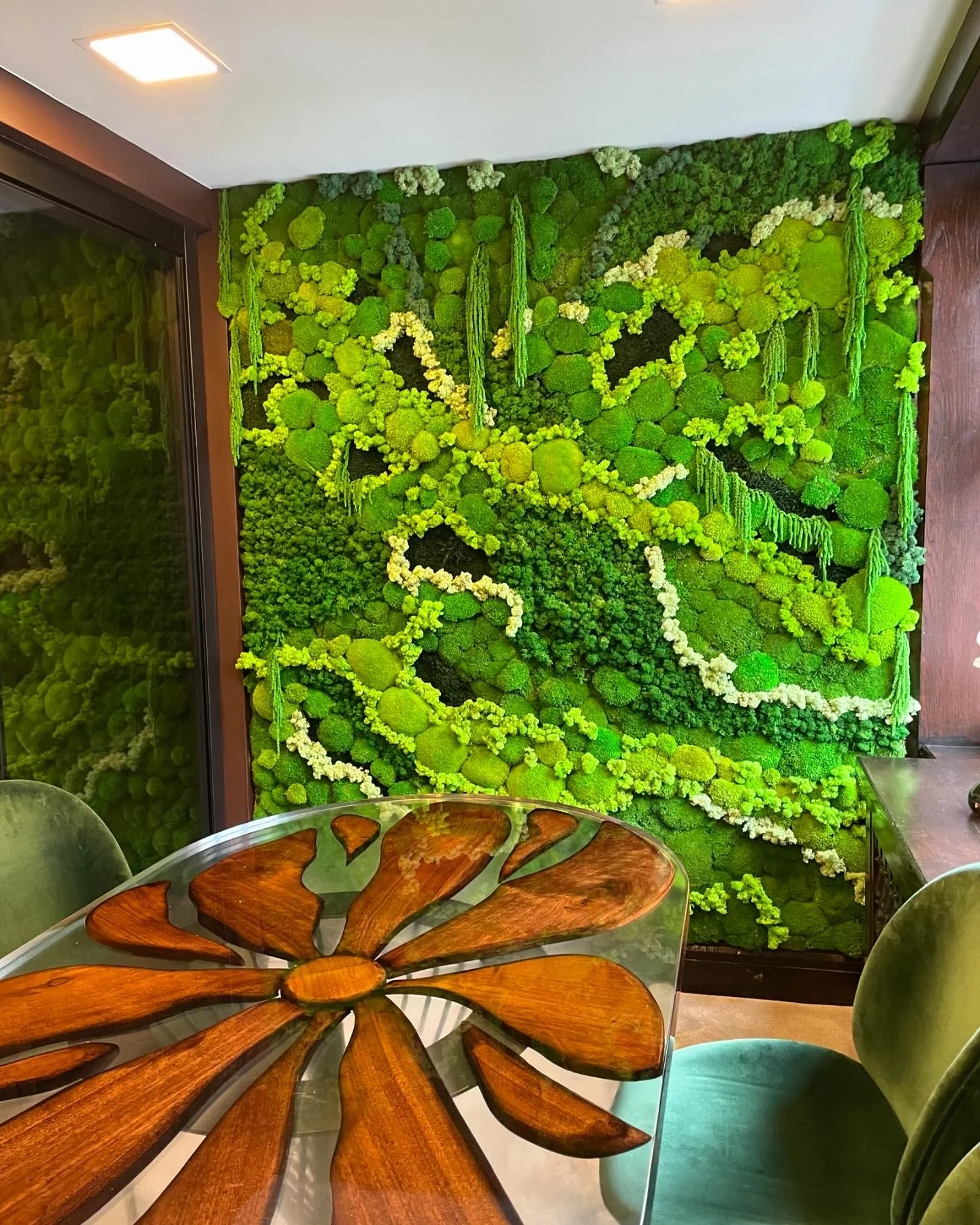 A green moss wall with various textured and shaped moss patches and small hanging moss or plants, next to a glass door. The image also shows a round table with a flower-shaped wooden top and green chairs around it.