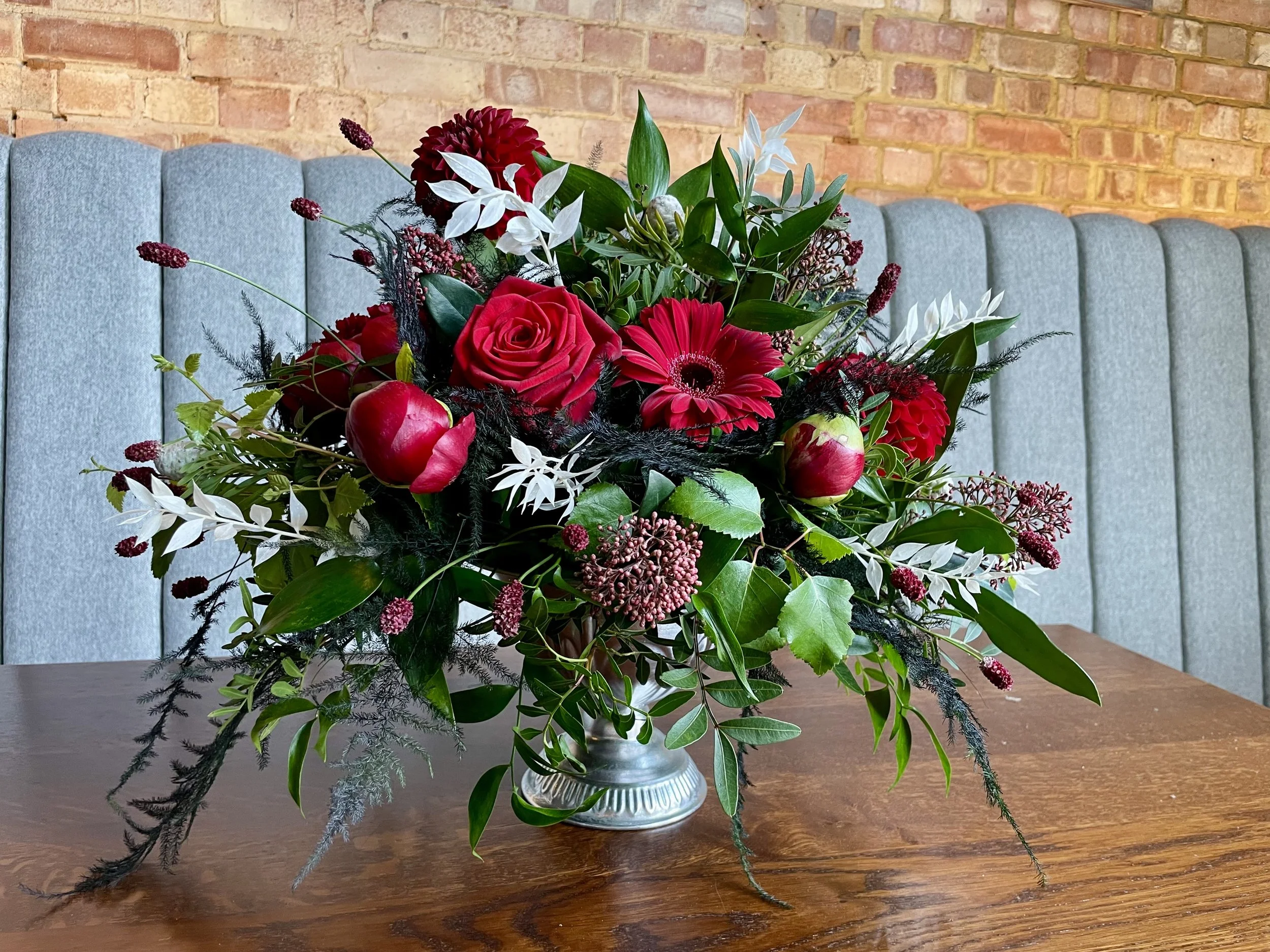 A floral arrangement in a silver vase placed on a wooden table, featuring red roses, white lilies, red gerbera daisies, greenery, and dark purple accent flowers, with a brick wall in the background.