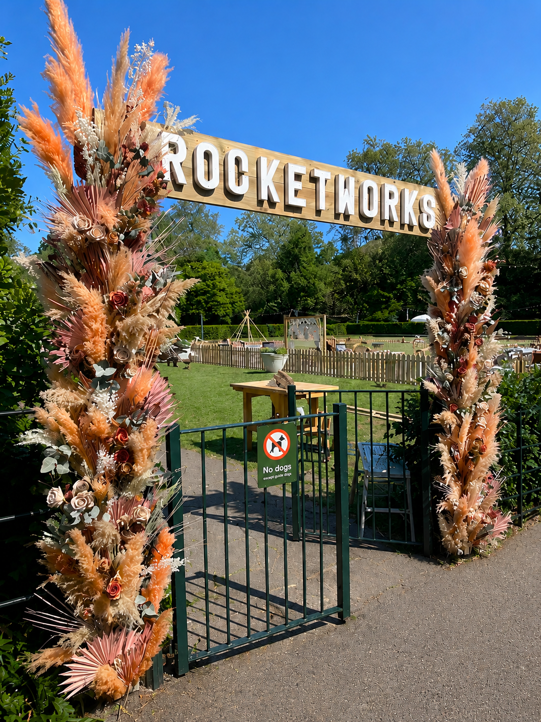 Decorated entrance gate for Rocketworks with floral arrangements on either side, a sign prohibiting dogs, and a grassy outdoor area with a wooden fence and trees in the background.
