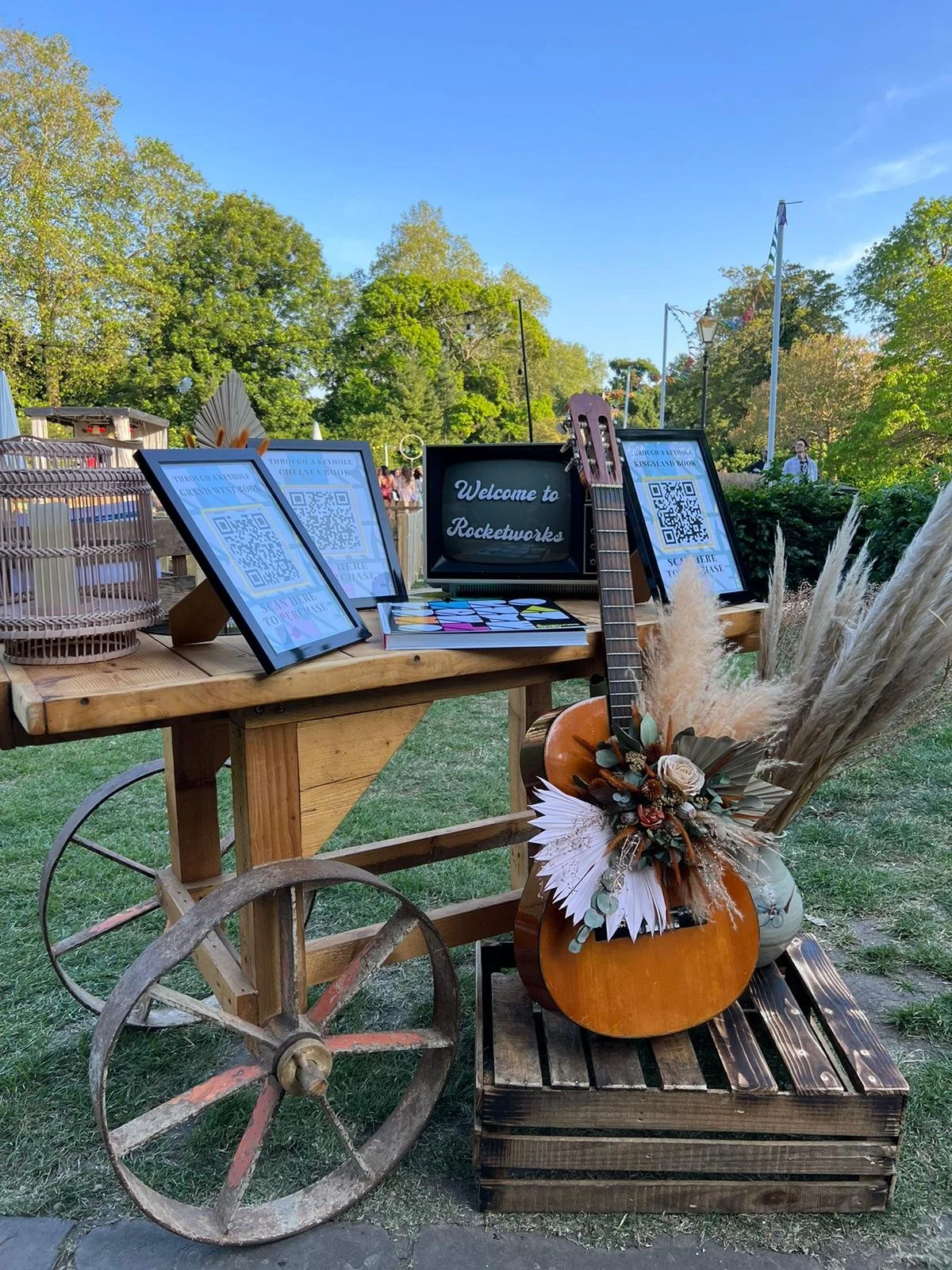 Outdoor event setup with a wooden table displaying framed signs with QR codes, a black sign reading "Welcome to Rocketworks," a stack of books, a guitar, and a large floral arrangement with pampas grass and a pumpkin, set against green trees and a blue sky.
