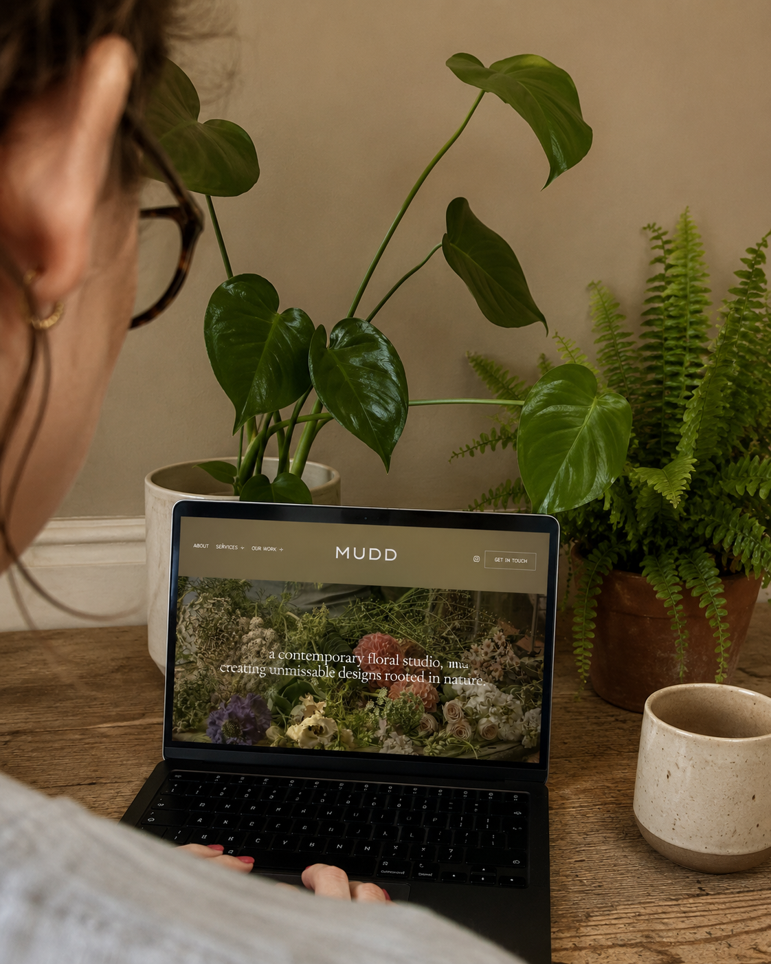 Person using a laptop at a wooden table, surrounded by green plants, including a large leafy plant in a white pot and a fern in a terracotta pot