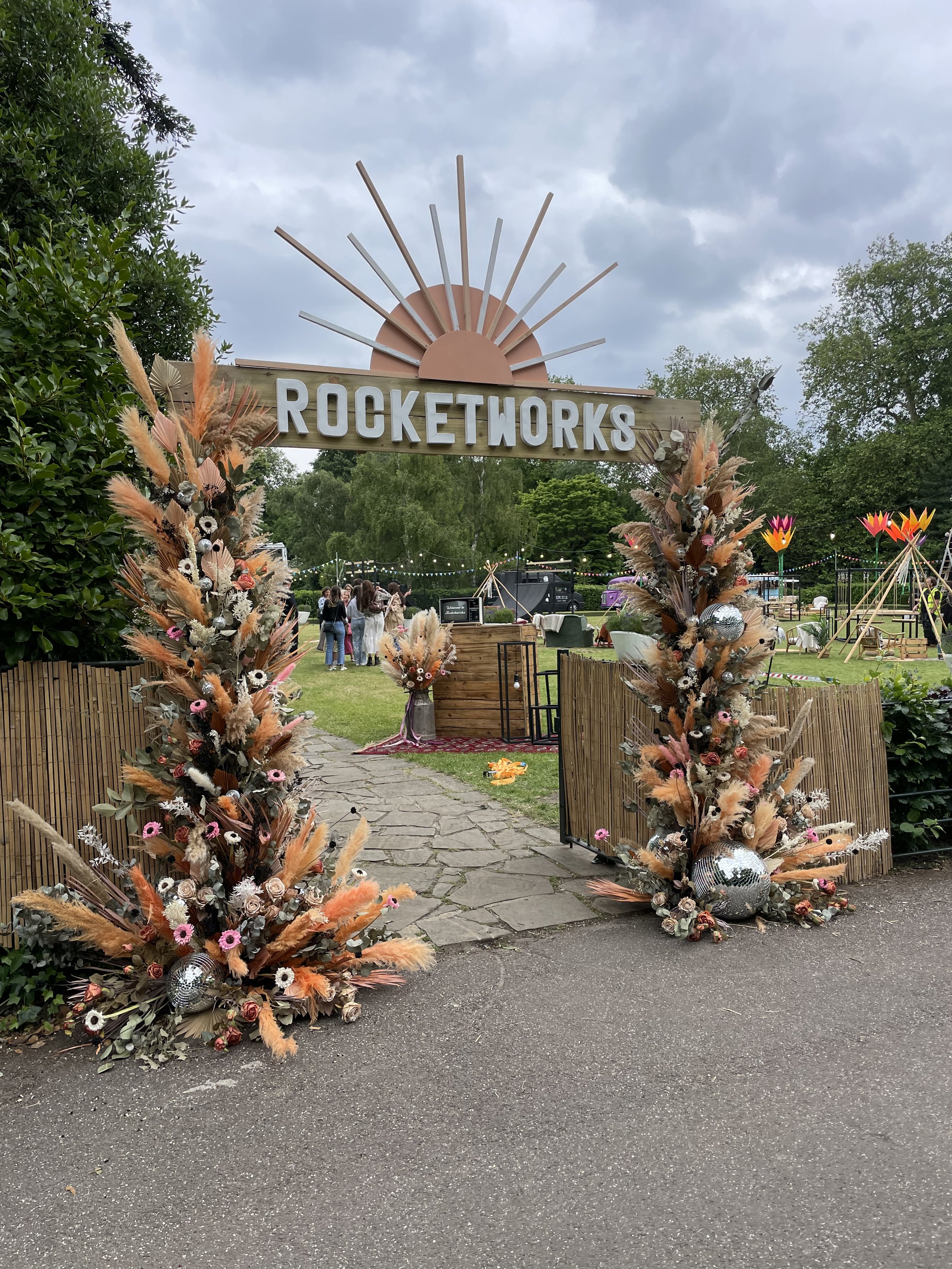 Decorative entrance with a wooden sign reading 'ROCKETWORKS' and floral arrangements made of dried flowers, pampas grass, and disco balls, leading into an outdoor event area under a cloudy sky.