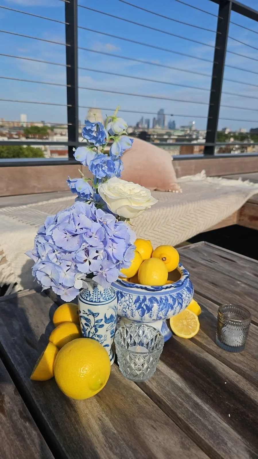 Vase with blue and white flowers and lemons on a wooden table outdoors with city skyline in the background.