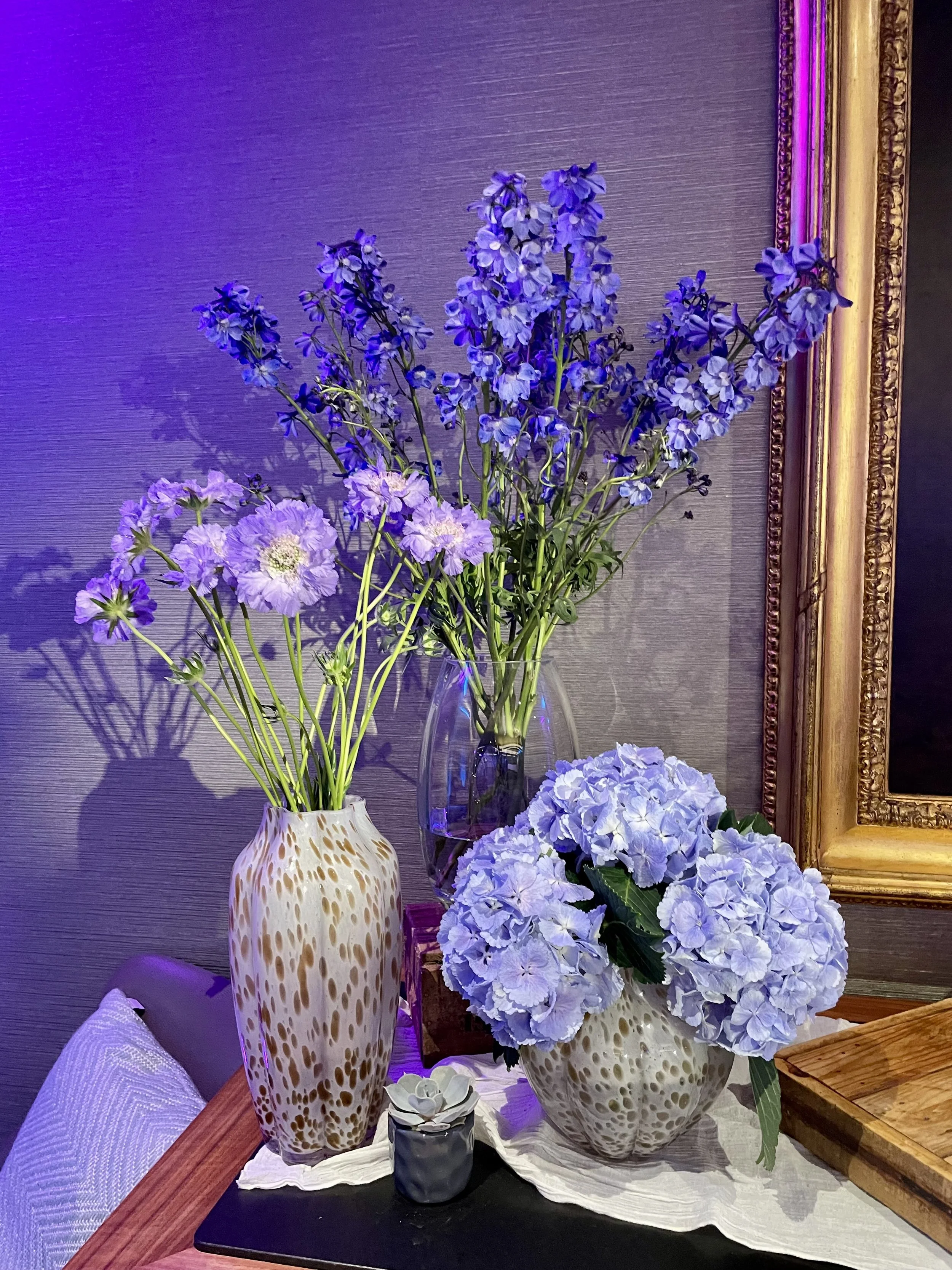 Arrangement of purple and white flowers in decorative vases on a table with a framed mirror or picture on the wall behind.