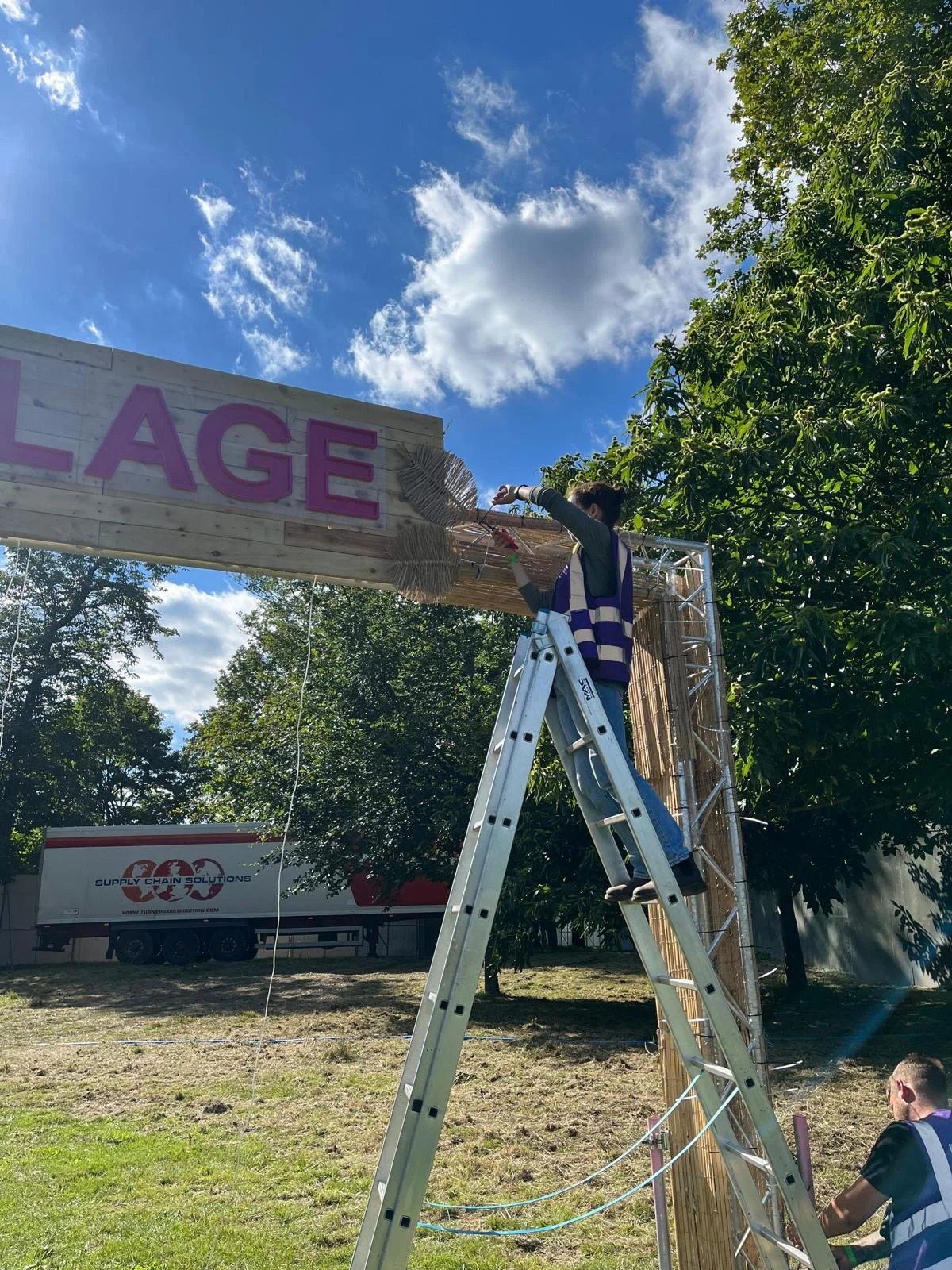 A person standing on a ladder is hanging or installing a purple sign with the letters "L AGE" outdoors during the daytime. The sky is blue with scattered clouds, and there are trees and a truck in the background.