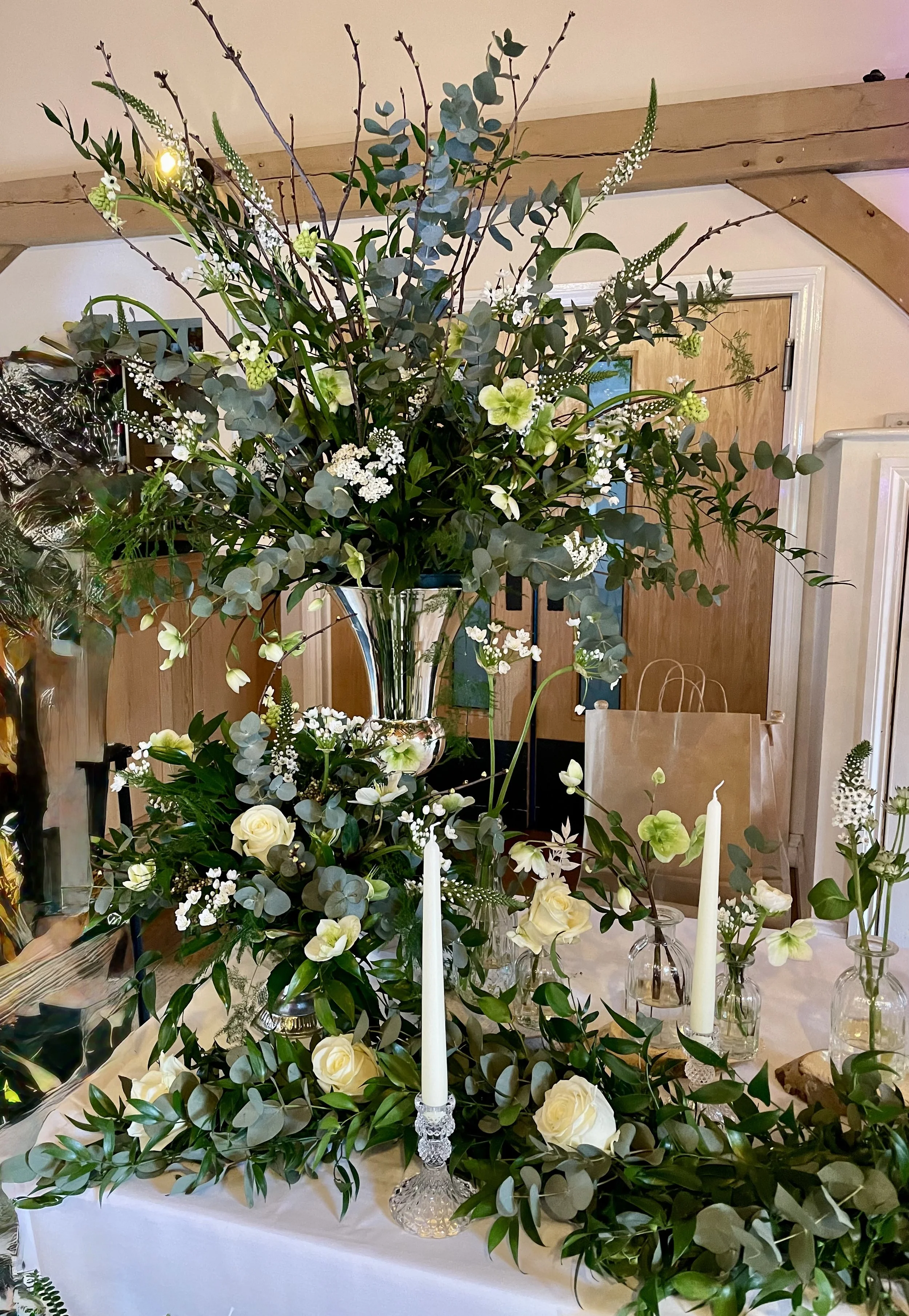 A floral arrangement with white roses, white and green flowers, and lush greenery in a silver vase, set on a table with white candles and glass vases, in a room with wooden beams and a wooden door in the background.