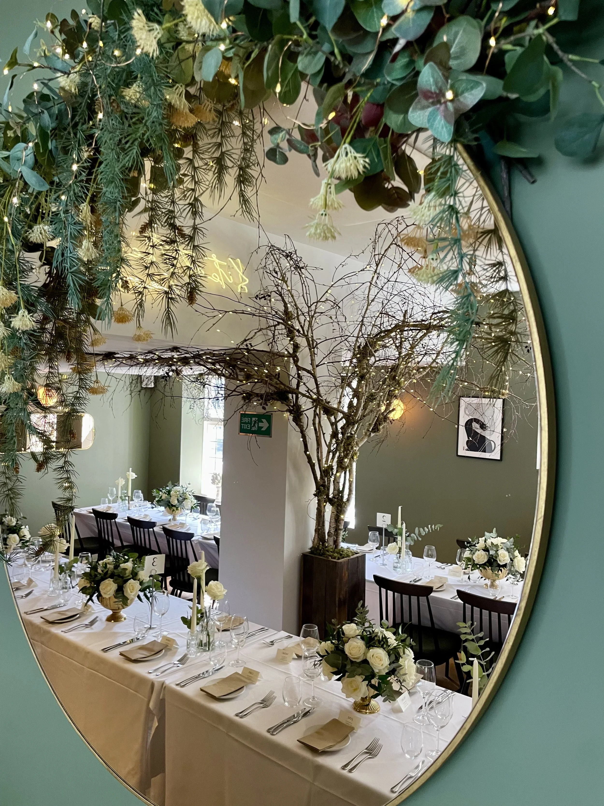 A decorated dining area with a large table set with white tablecloths, flowers, candles, and glassware, viewed through a mirror with a green frame that has hanging plants and lights.