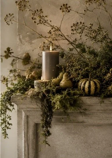 Decorative arrangement on a fireplace mantle with a gray candle, dried branches, greenery, and small decorative gourds.