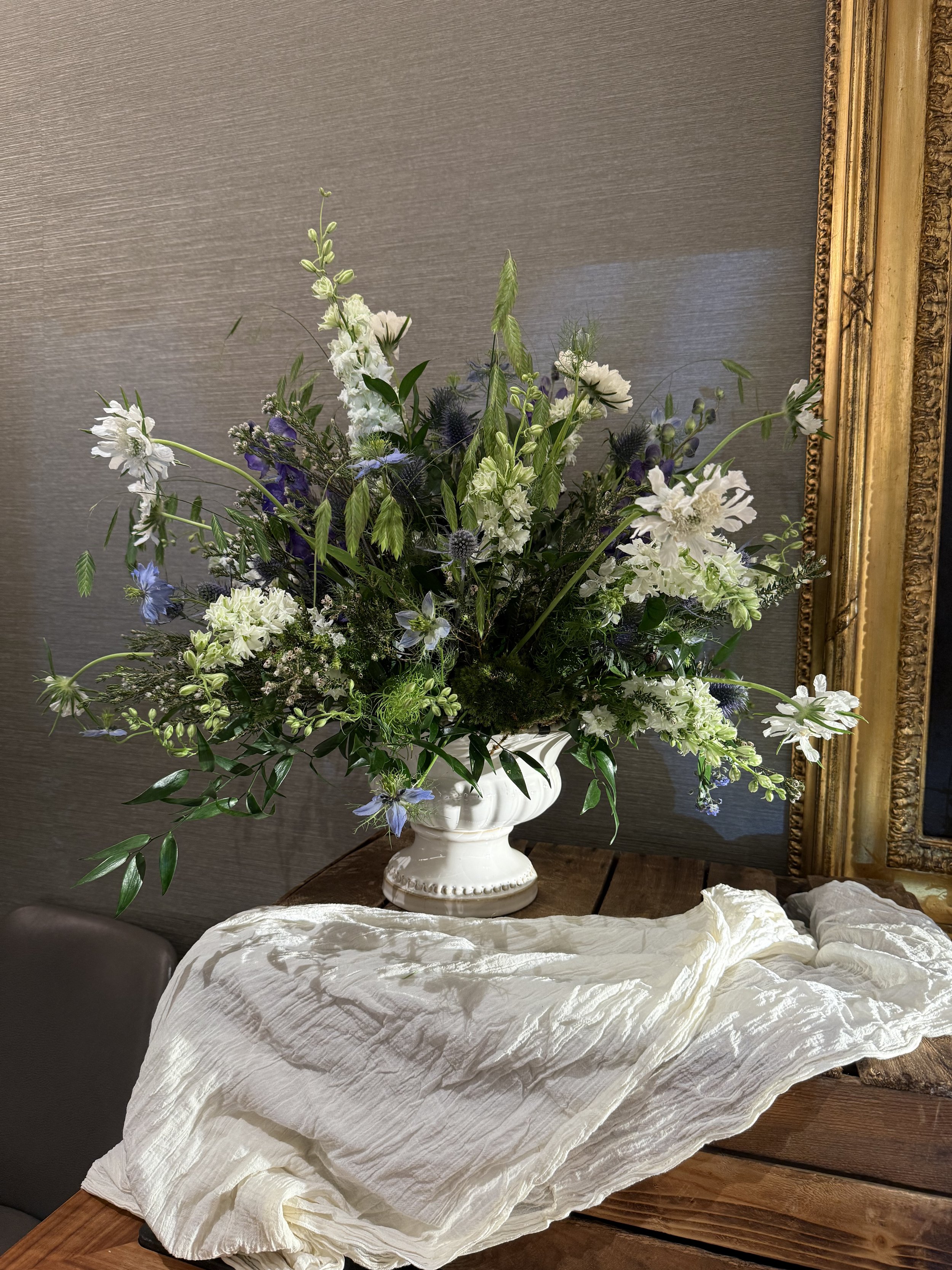 A large arrangement of white and purple flowers in a white ceramic vase on a wooden table, with a textured grey wall and gold-framed mirror in the background.