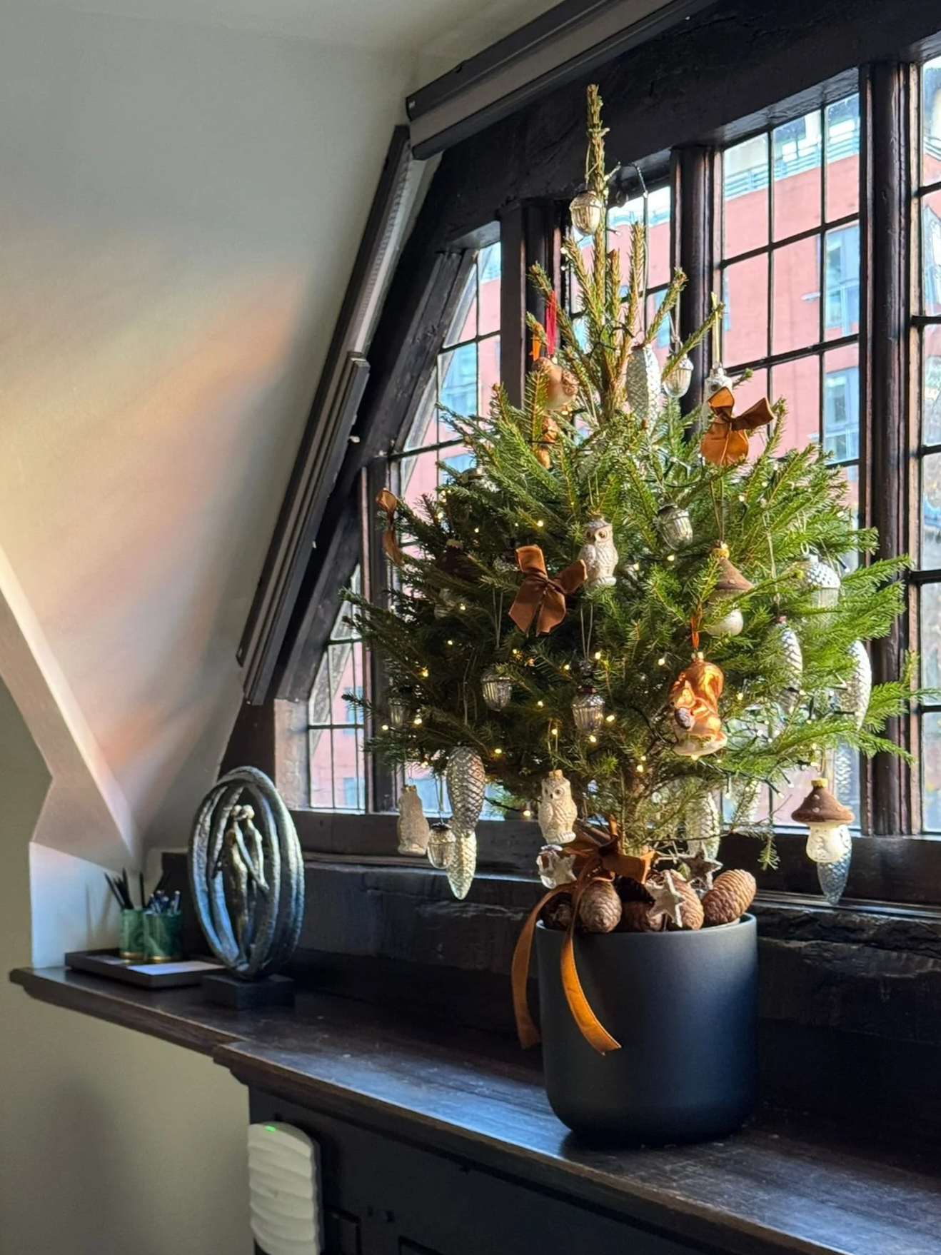Small decorated Christmas tree in a black pot on a wooden window sill, with ornaments, lights, and ribbons, in front of a large window with black framing, with a brick building outside.