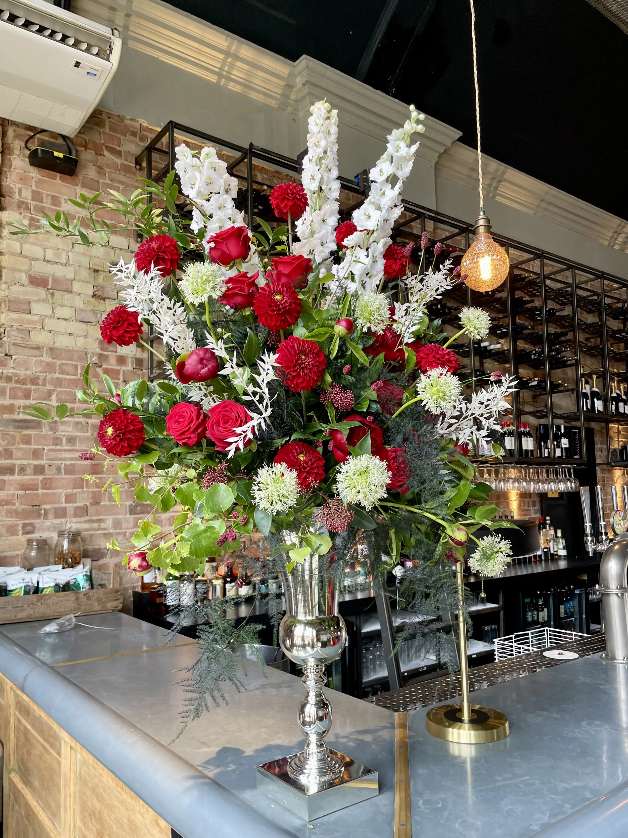 A large floral arrangement with red roses, red dahlias, white gladiolus, and white spider mums in a silver vase on a bar counter. Behind the flowers is a wine rack in a brick wall, along with shelves of wine bottles and glasses. A hanging light fixture glowing warmly is also visible.