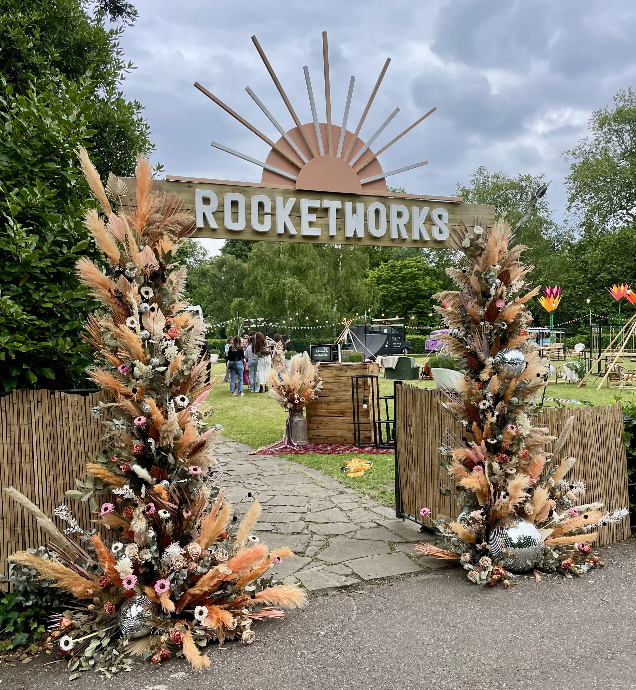 Decorative archway with a sign reading 'ROCKETWORKS' at the entrance of an outdoor event, surrounded by floral arrangements and greenery, with people gathering in the background under a cloudy sky.