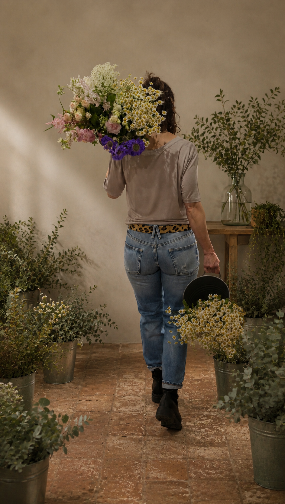 Woman walking through a flower shop with a large bouquet of flowers covering her face, surrounded by potted plants and flowers, holding a bucket of daisies.