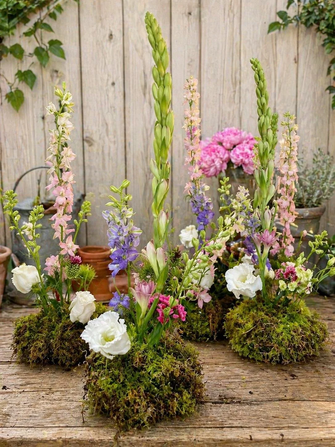 Arrangement of blooming flowers including pink, purple, and white varieties on a wooden surface with potted plants and a wooden fence in the background.