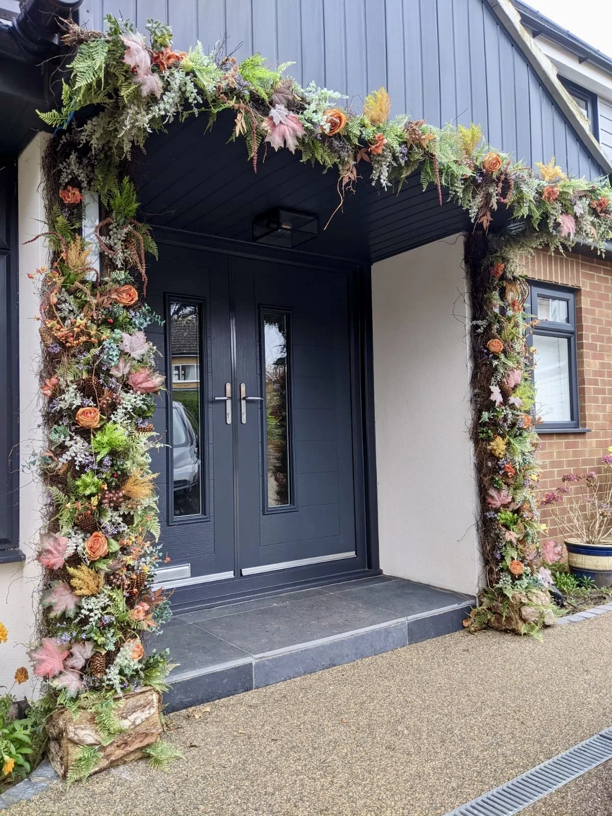 Front entrance of a house decorated with a large floral arrangement surrounding the door and framing the doorway, featuring a mix of greenery, ferns, orange roses, and other colorful flowers.