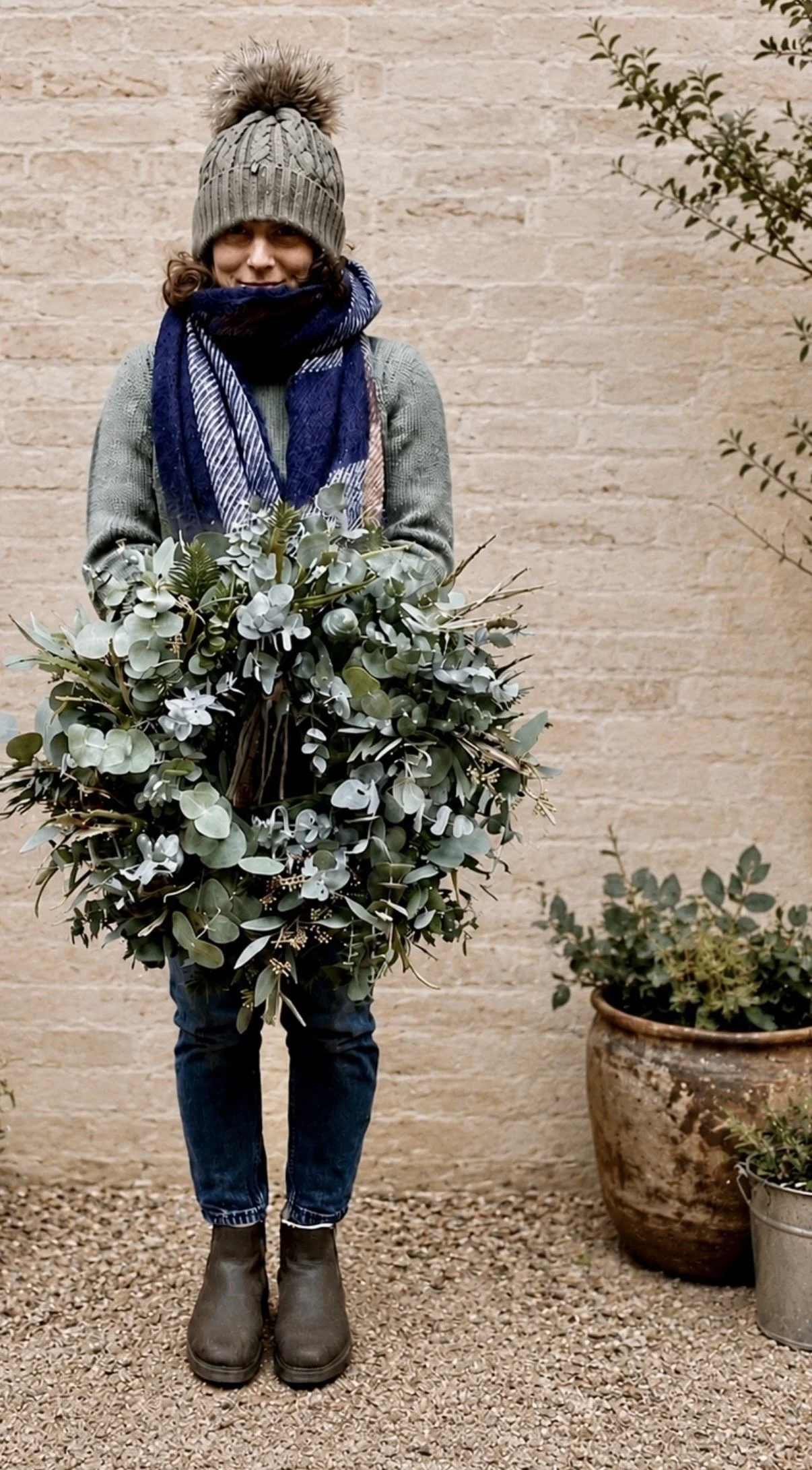 Person wearing a gray knit hat with a pom-pom, a blue scarf, a greenish sweater, dark jeans, and brown boots standing outdoors in front of a beige brick wall, holding a large bouquet of green-leafed plants.