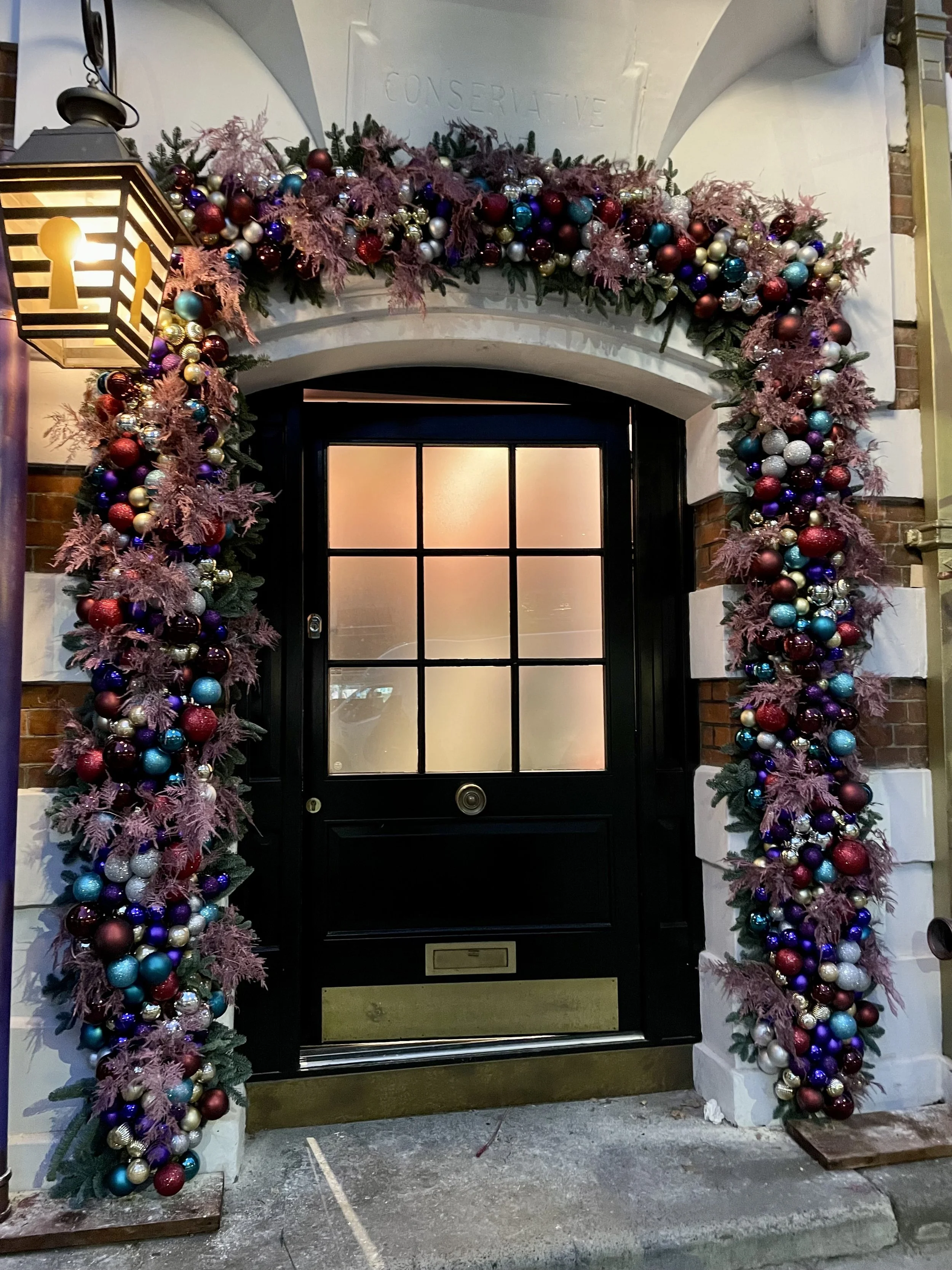 Decorated archway with pink, red, blue, silver, and purple Christmas ornaments and greenery at a building entrance, with a black door behind.