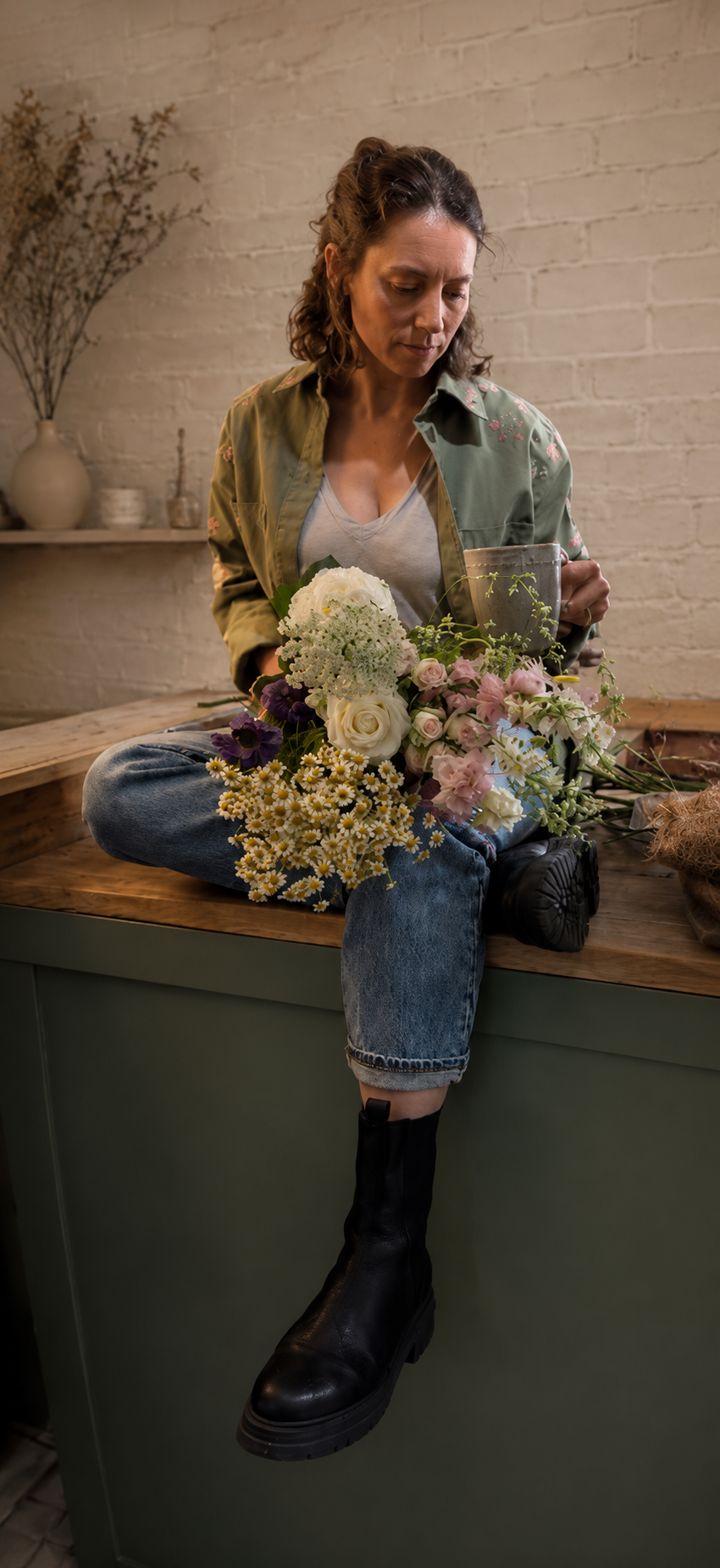 Woman sitting on a wooden counter with one leg crossed, holding a mug in one hand and a bouquet of flowers in her lap, wearing a green jacket, gray shirt, and black boots.