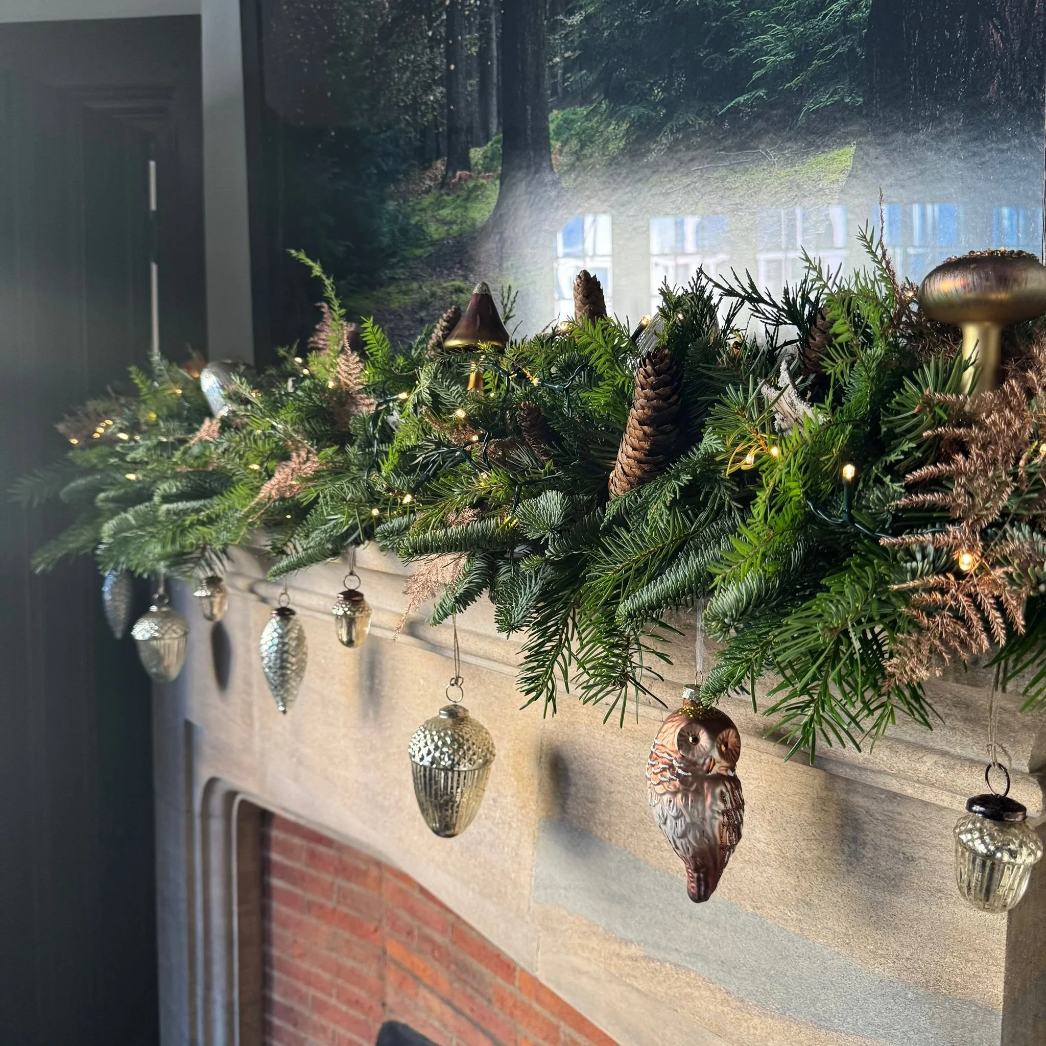 Green Christmas fire place arrangement with glass pine cone decorations, woodland animal themed glass decorations and small fairy lights. Placed on a beige mantle surface near a brick wall, with a blurry window in the background.