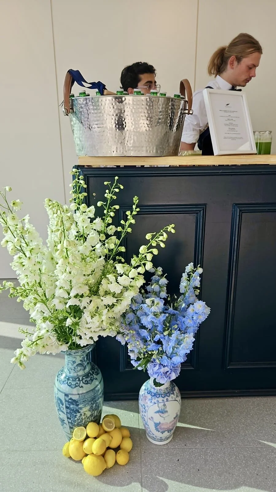 A flower arrangement with white and blue flowers in decorative vases, a pile of lemons, and a reception counter with two people, one of whom is a woman in a white shirt with a black bow tie, and a large metal bucket with bottles on top.