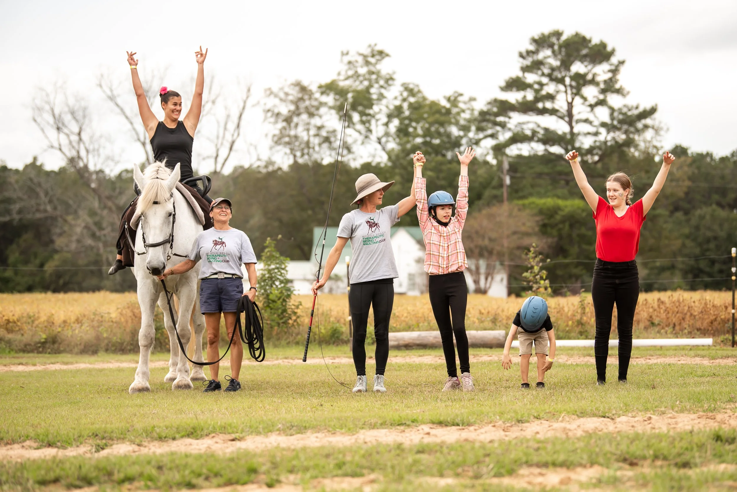 Group of people celebrating outdoors on a field, with a woman on horseback in the center raising her arms, others around her cheering, and a child with a helmet bending down.